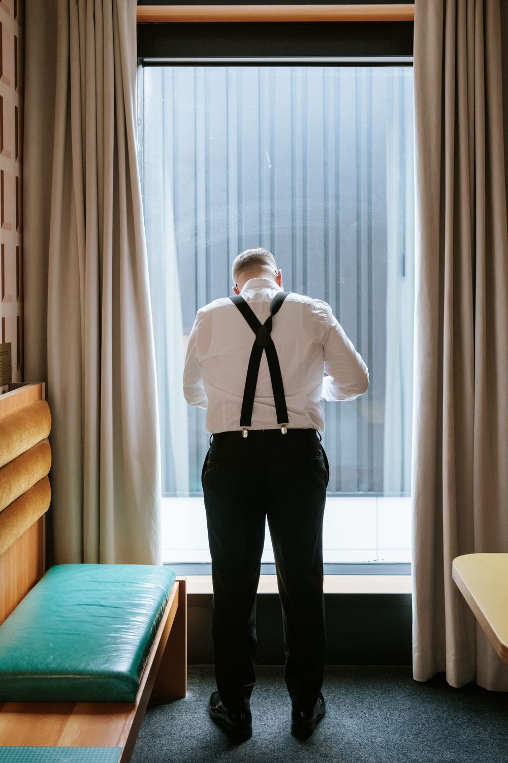 Person in formal attire with suspenders looking out window in hotel room.