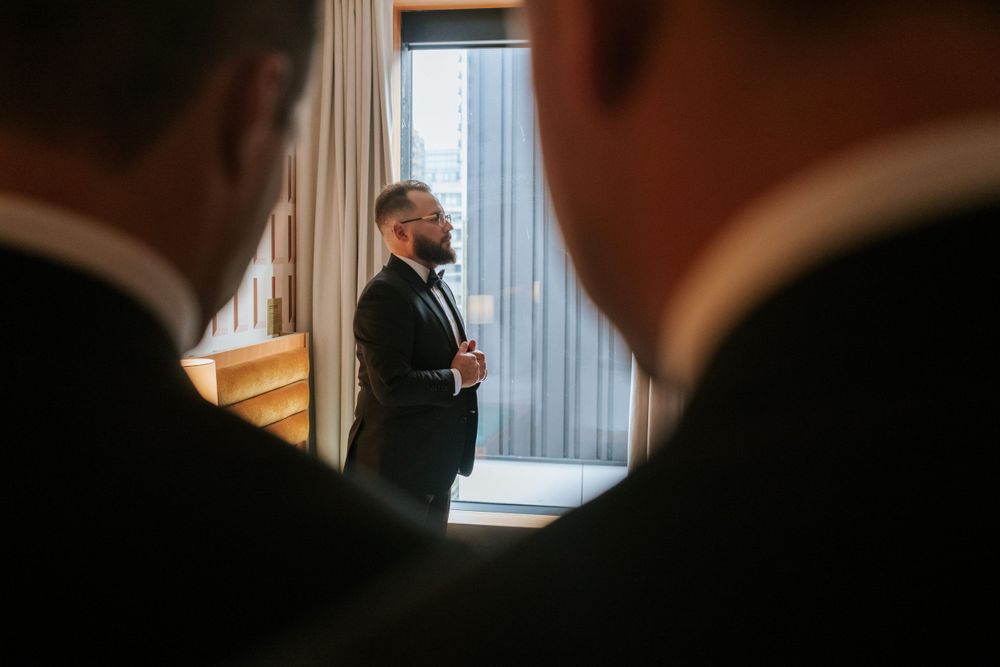 Wedding day preparation scene through silhouettes looking at suited figure by window.