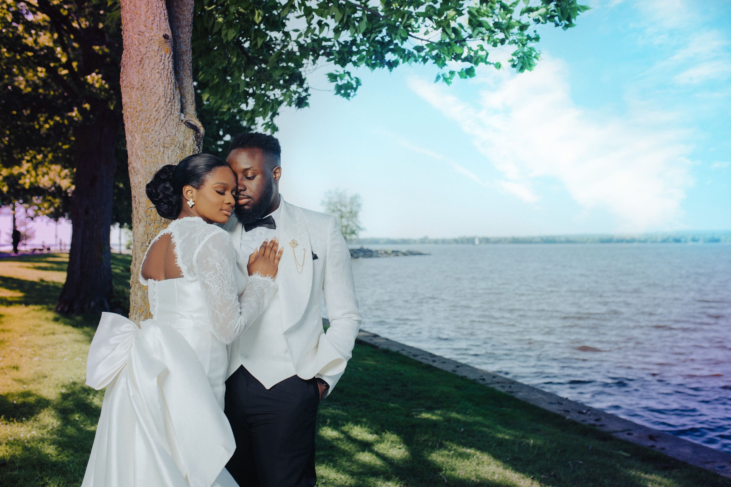 A romantic outdoor wedding photo captures a couple embracing by the waterfront on a sunny day.