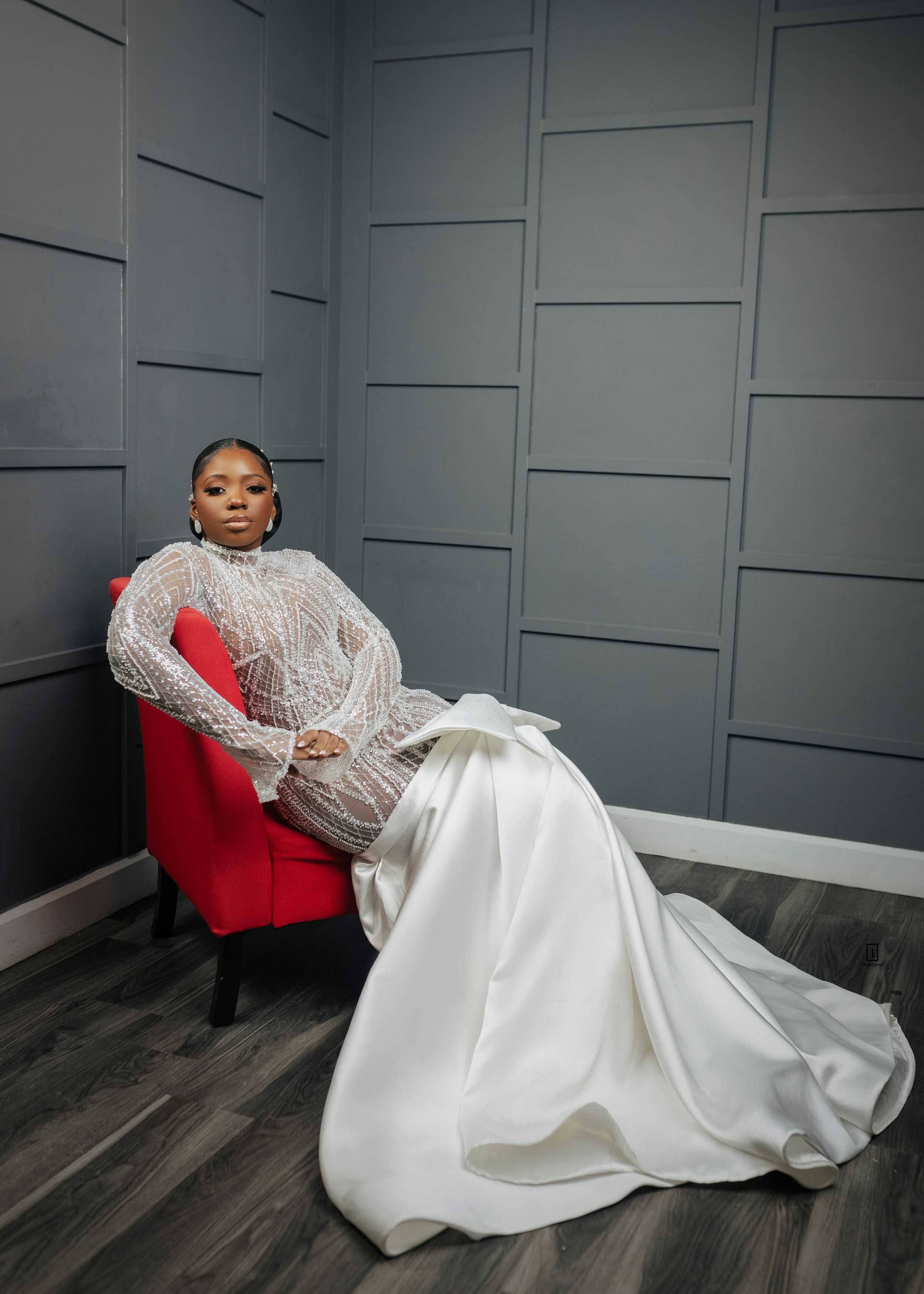 An elegant wedding gown with lace sleeves and flowing white satin skirt is displayed on a red velvet chair against a gray wall.