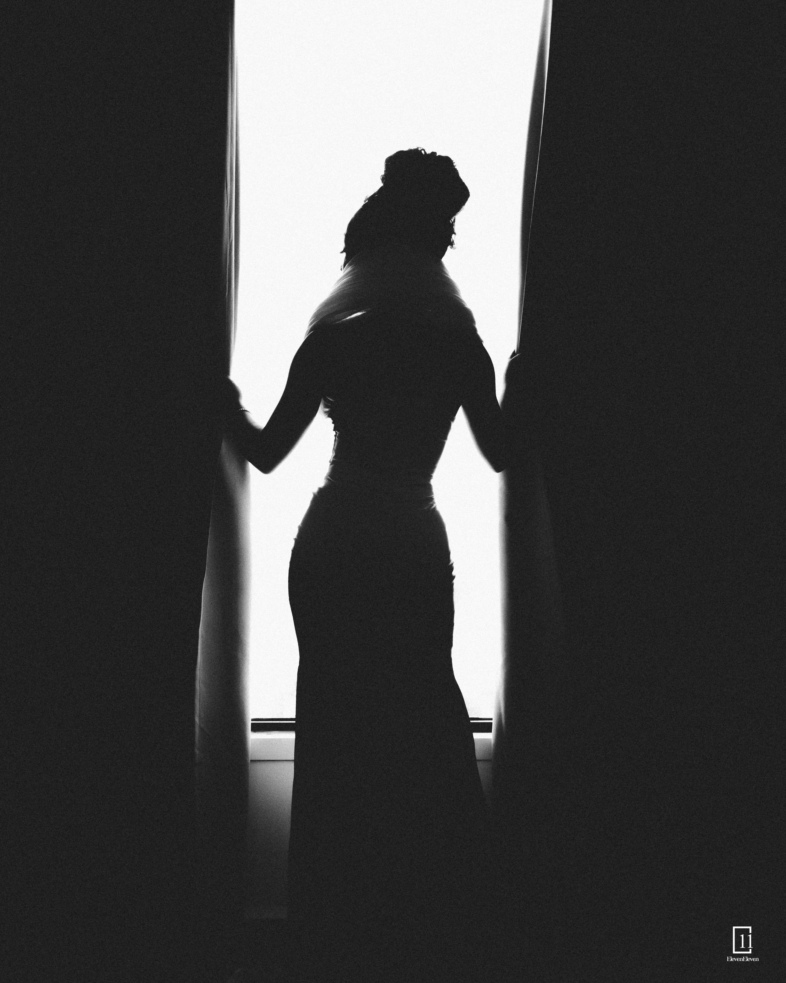 Dramatic black and white silhouette of a woman in an elegant dress standing between curtains by a window.