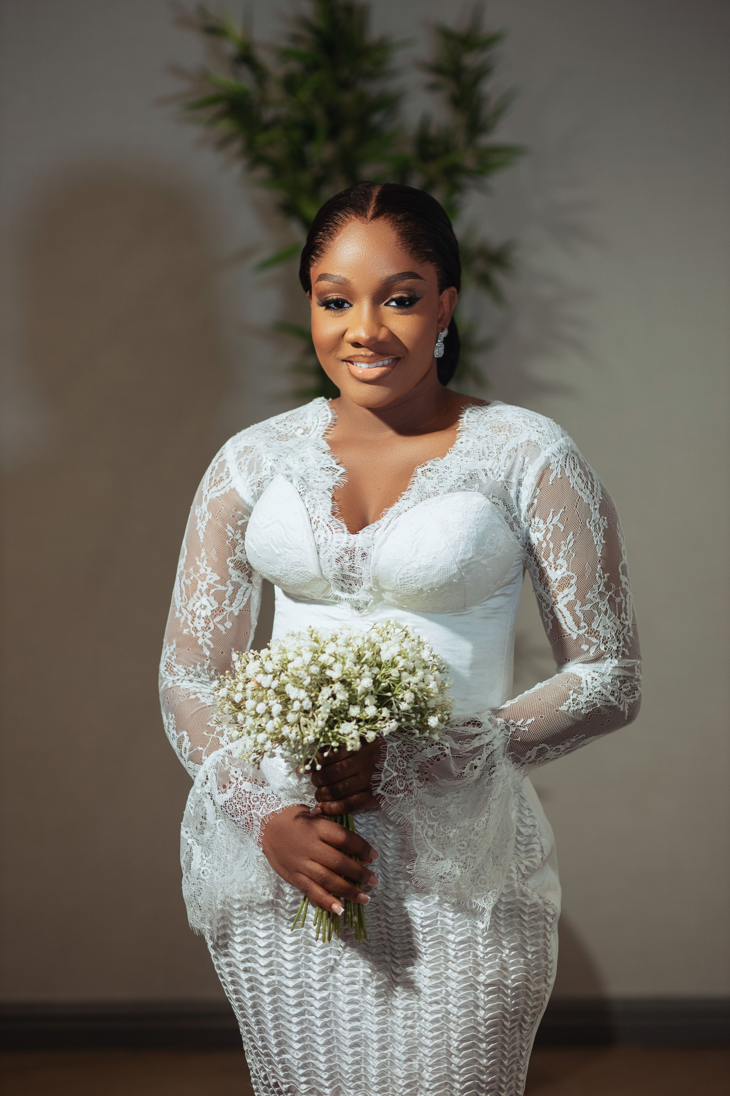 A bride poses in an elegant white lace wedding gown with long sleeves and holds a baby's breath bouquet.