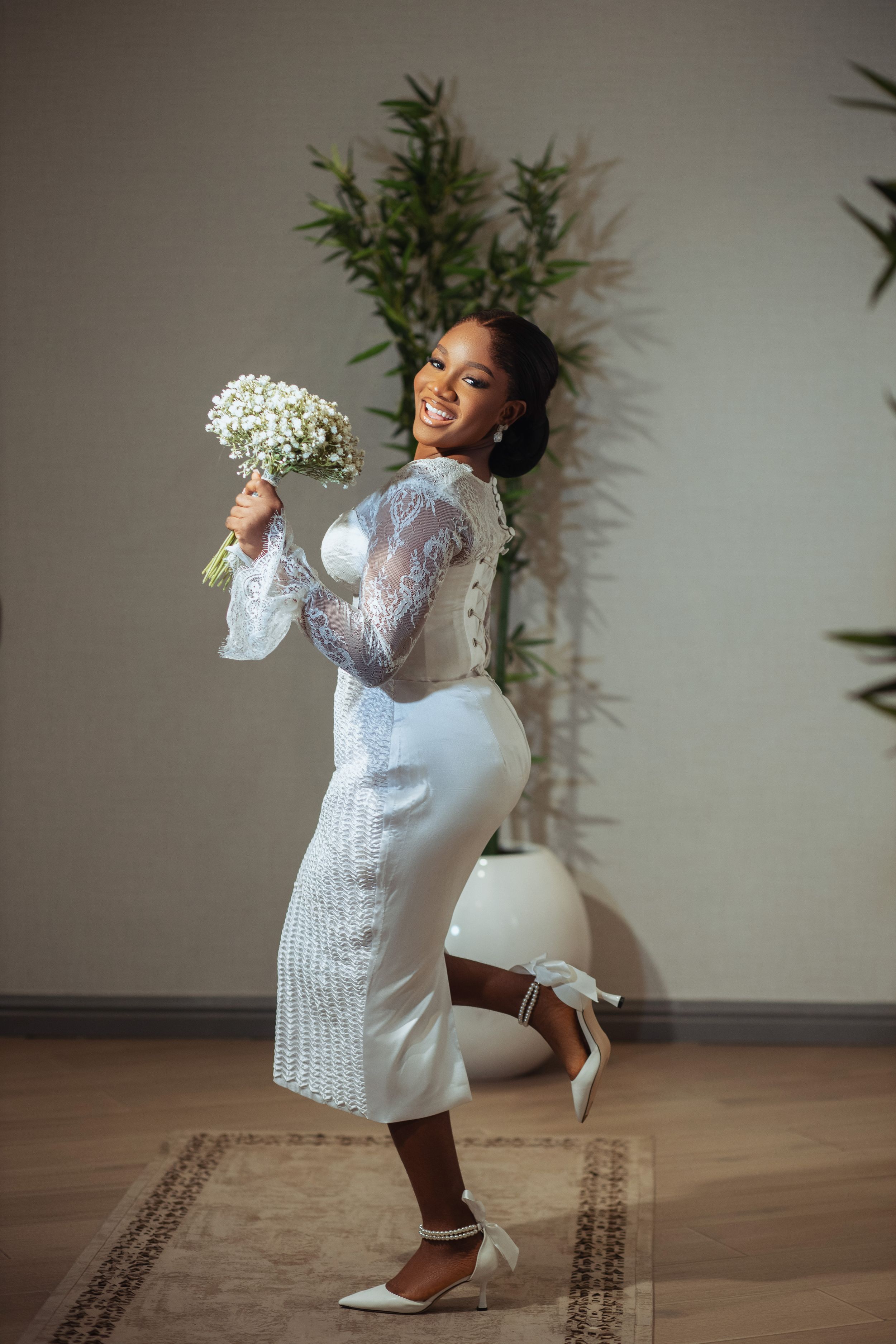 A joyful bride in a fitted white lace dress playfully poses with her bouquet against greenery.