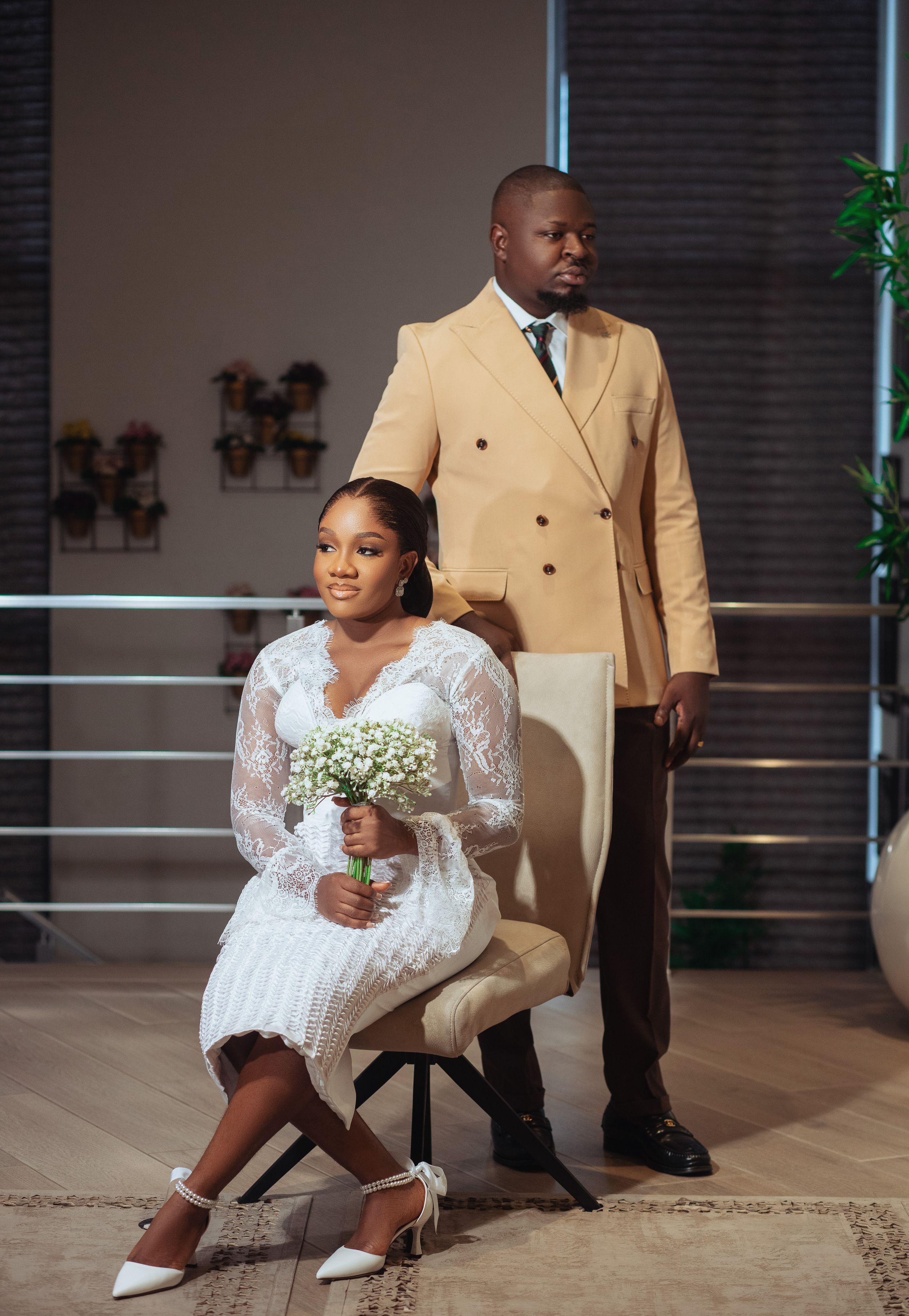 Wedding portrait with bride seated in white dress and groom standing in tan suit.
