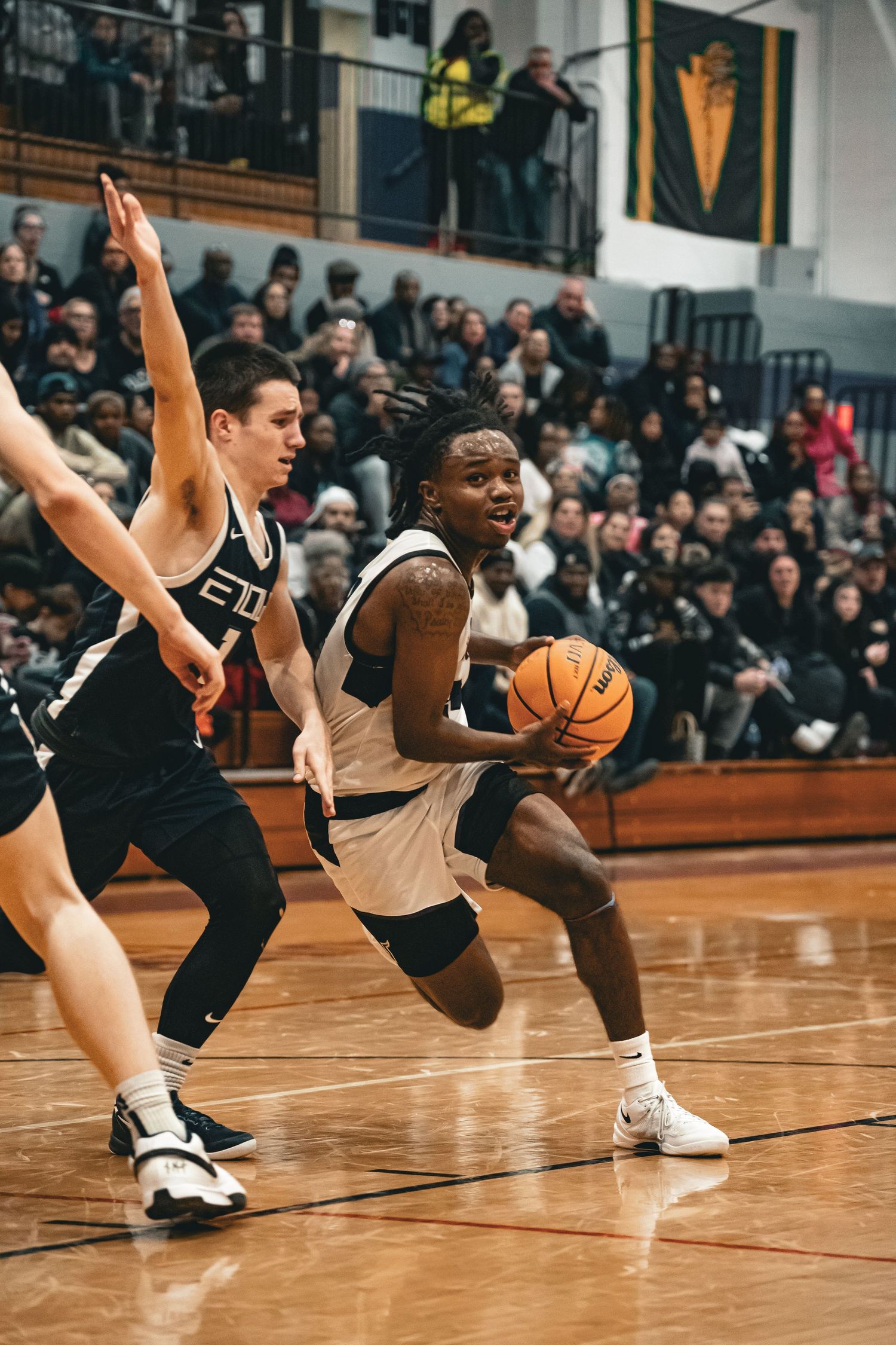 Two basketball players compete during an intense game on a wood court with spectators watching from packed bleachers.
