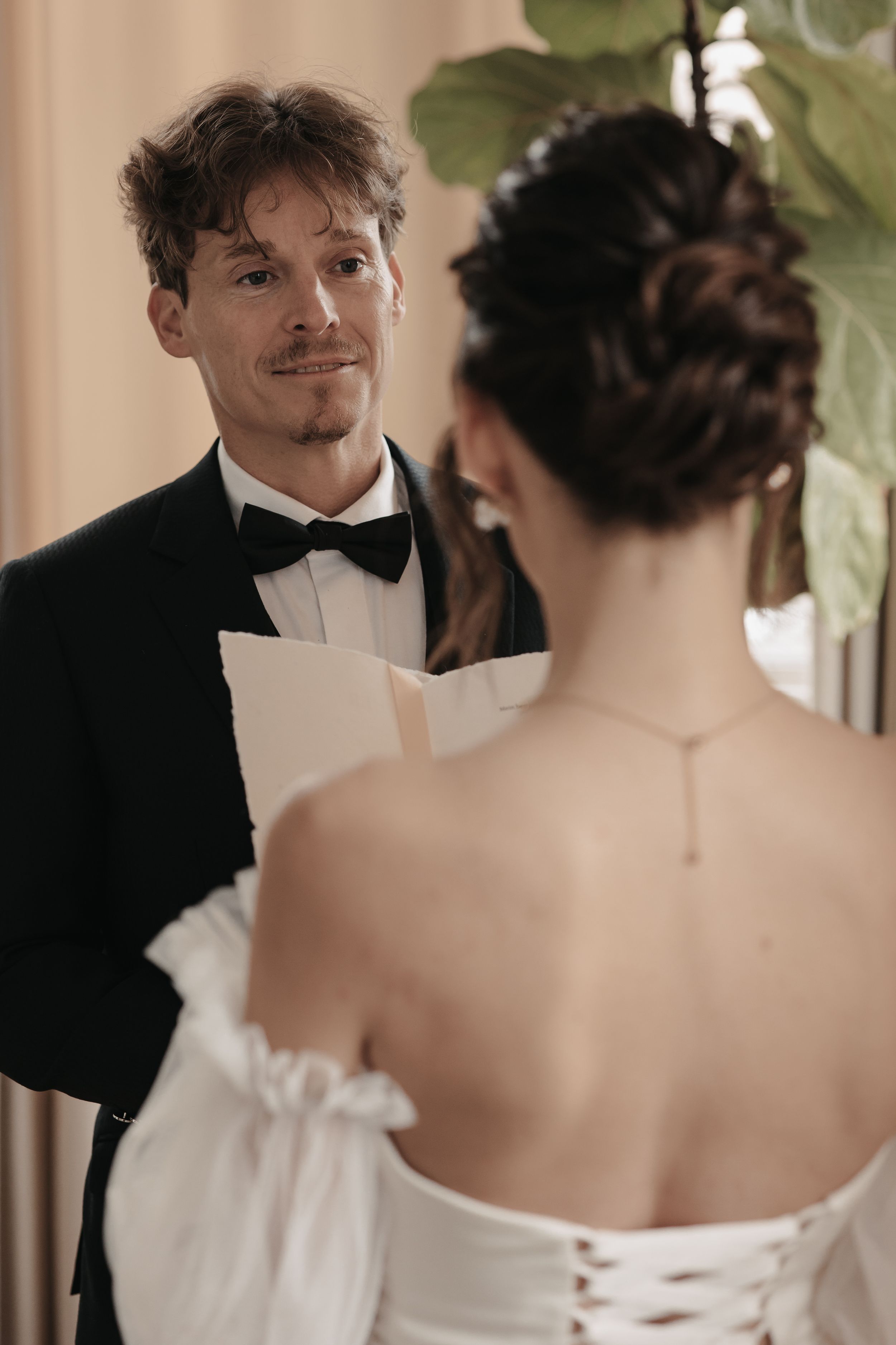 A groom in a black tuxedo reads wedding vows while facing the bride in her white corseted dress.