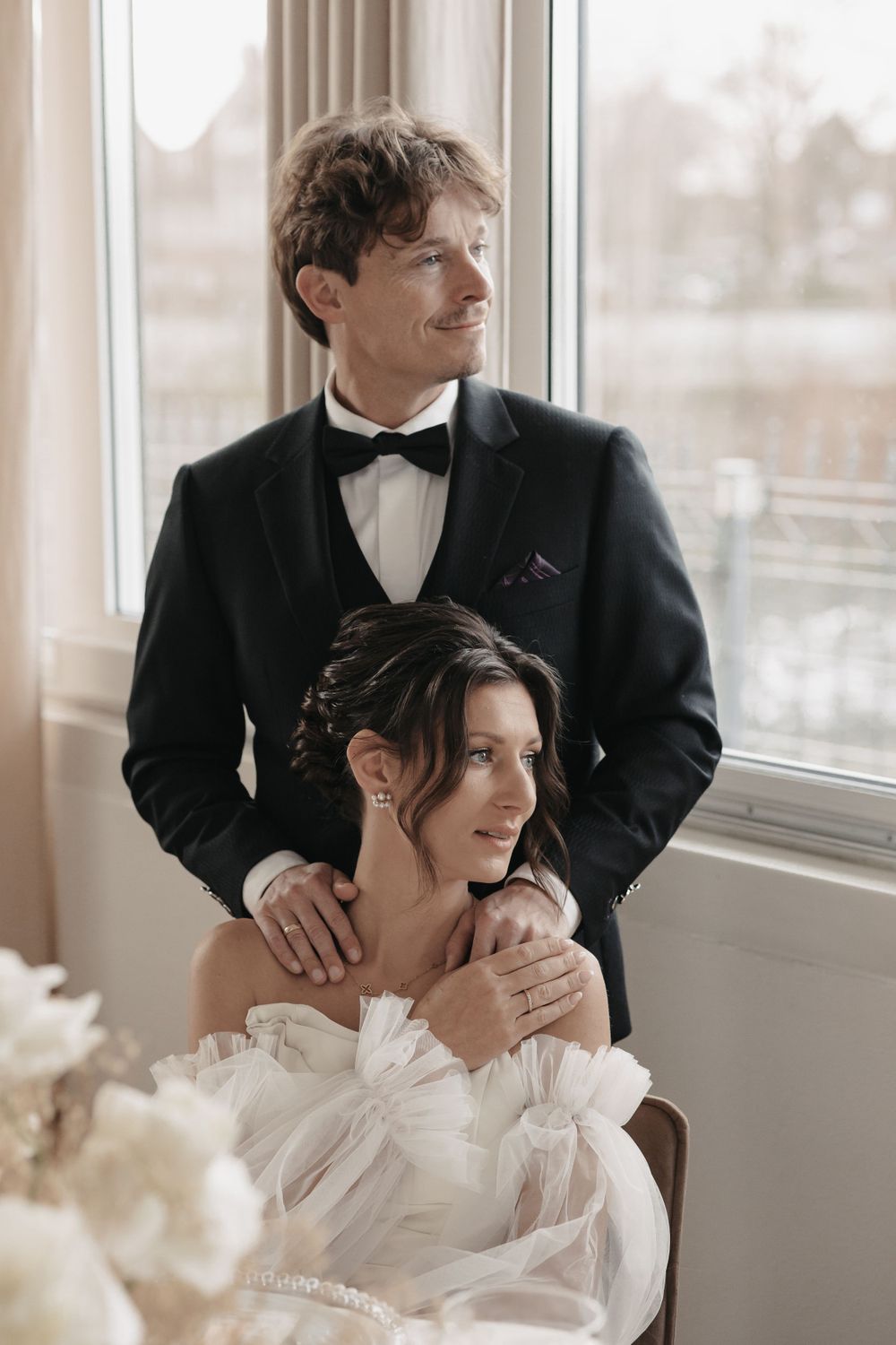 Elegant couple in formal attire pose near window, with bride in ruffled white dress and groom in classic black tuxedo.