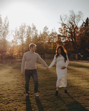 A couple holds hands during golden hour in a field with autumn trees in the background.