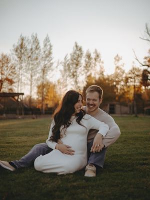 A couple sits together on grass during golden hour in a scenic outdoor setting.