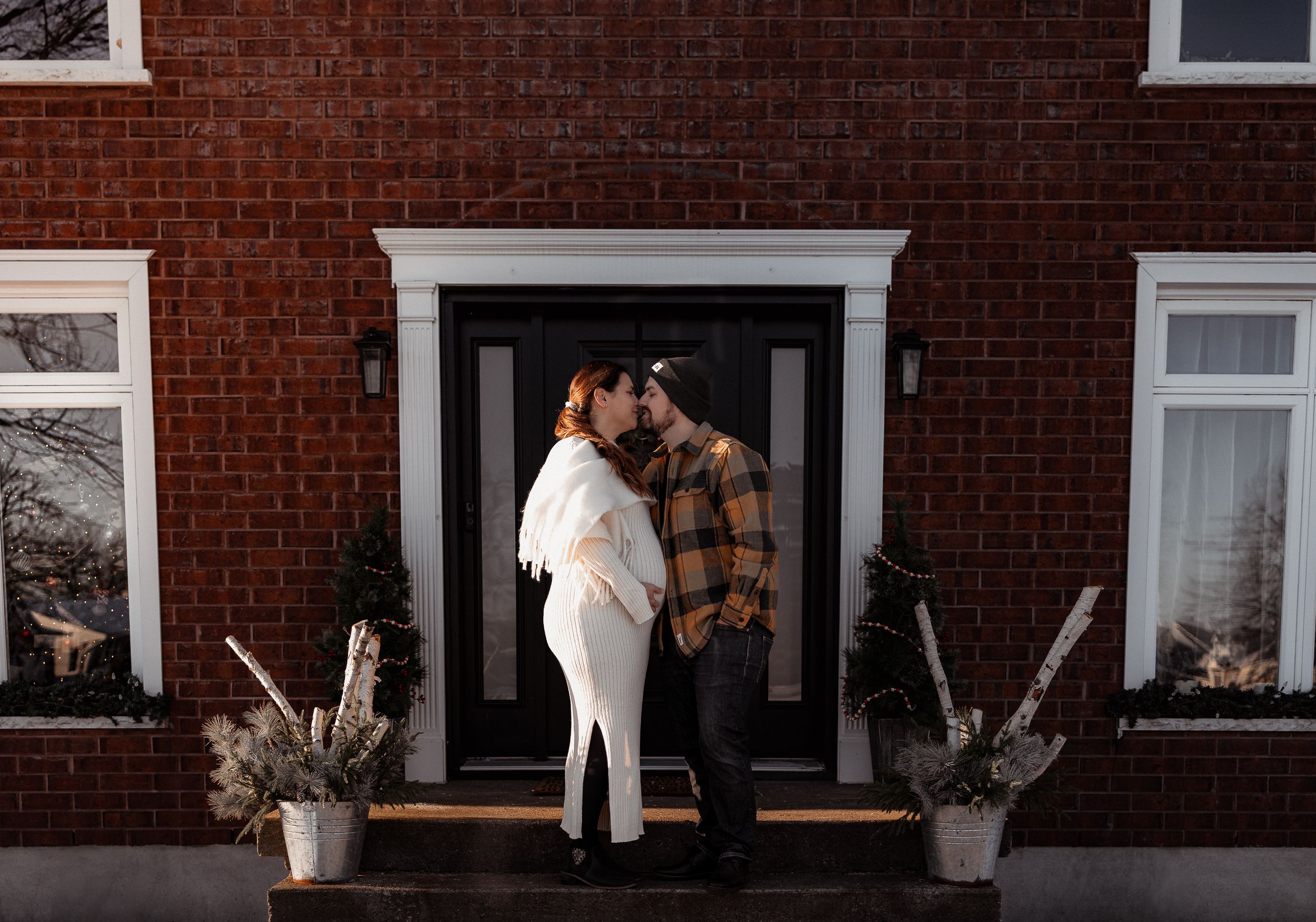 A cozy winter scene shows a couple embracing on their front porch of a brick house.