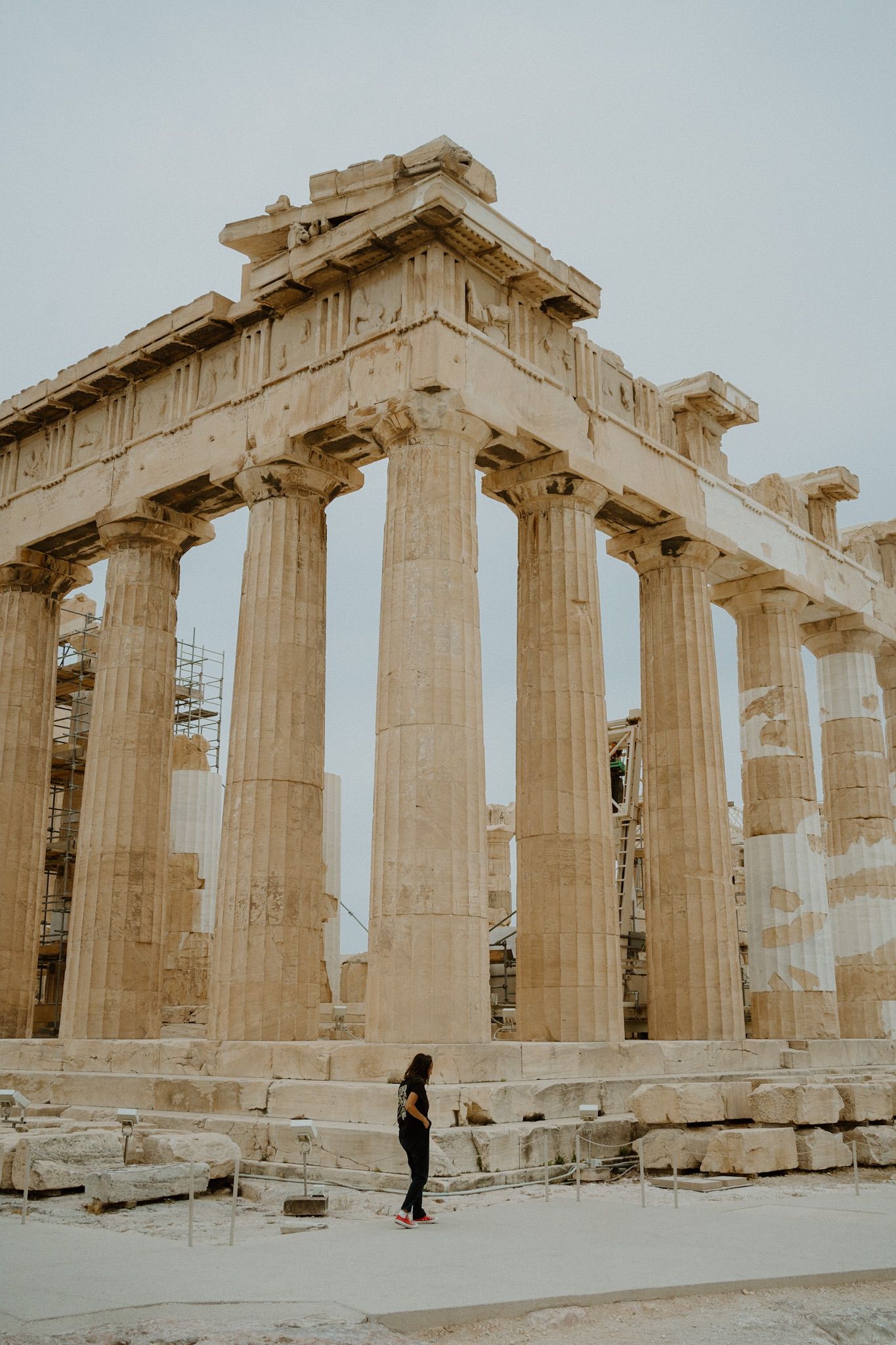 A lone figure stands beneath the towering ancient columns of the Parthenon at the Acropolis in Athens.