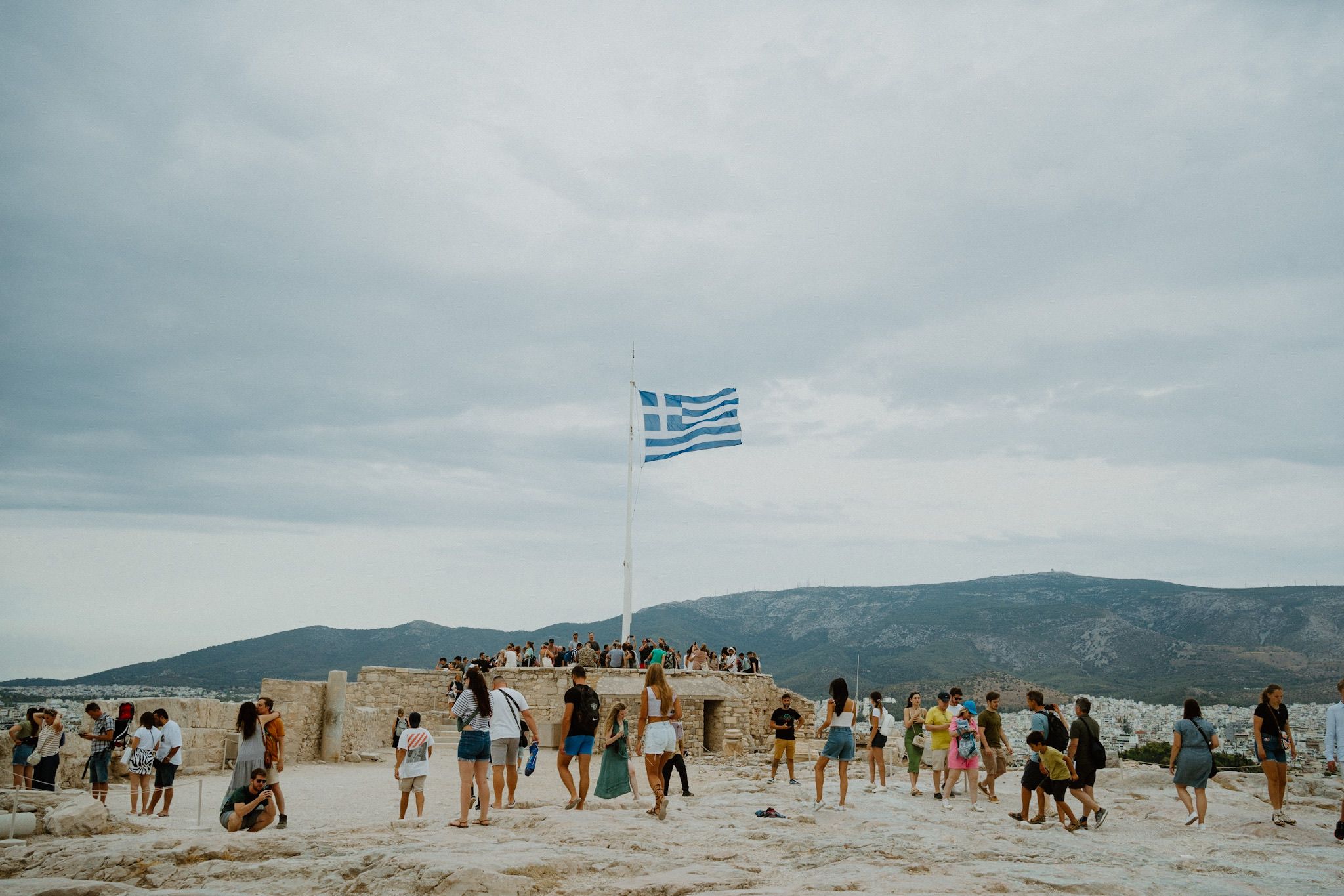 Large group of tourists gather at hilltop viewpoint with Greek flag waving against cloudy sky and mountain backdrop.