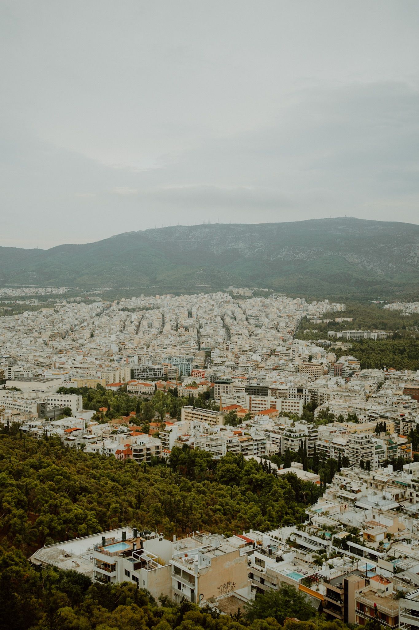 Aerial view of Athens cityscape with densely packed buildings against mountain backdrop under cloudy skies.
