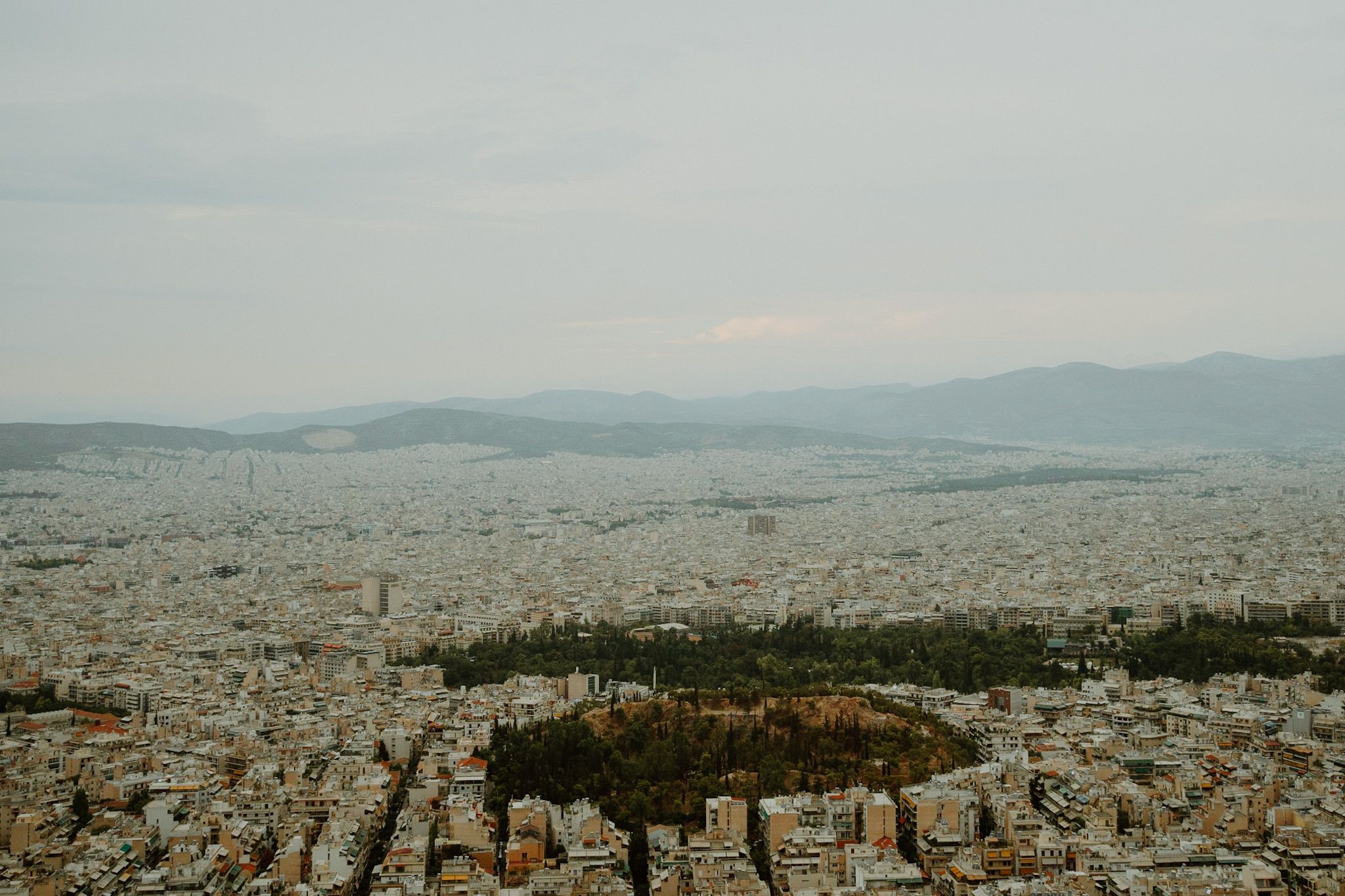 Aerial view of Athens cityscape with dense urban buildings stretching to distant mountains under an overcast sky.