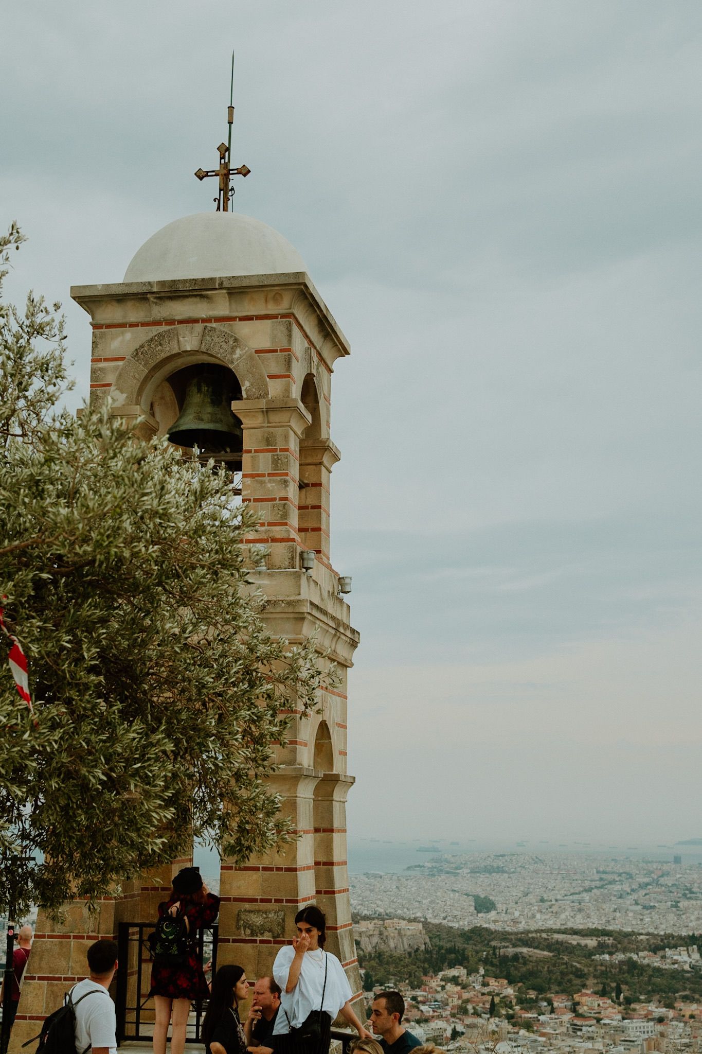 Historic bell tower overlooking Athens cityscape with visitors gathering at Mount Lycabettus on a cloudy day.