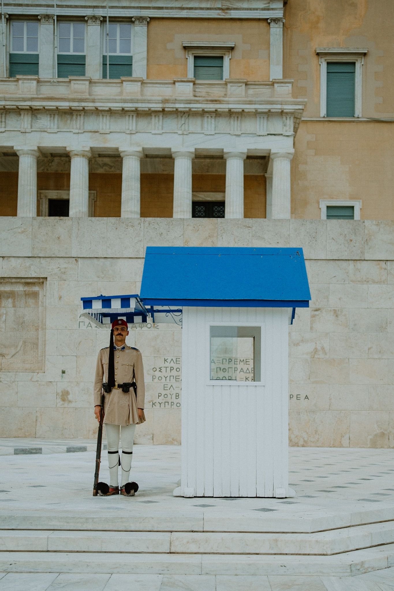 A traditional guard stands at attention next to a white guard booth with blue roof in front of a classical building in Athens.