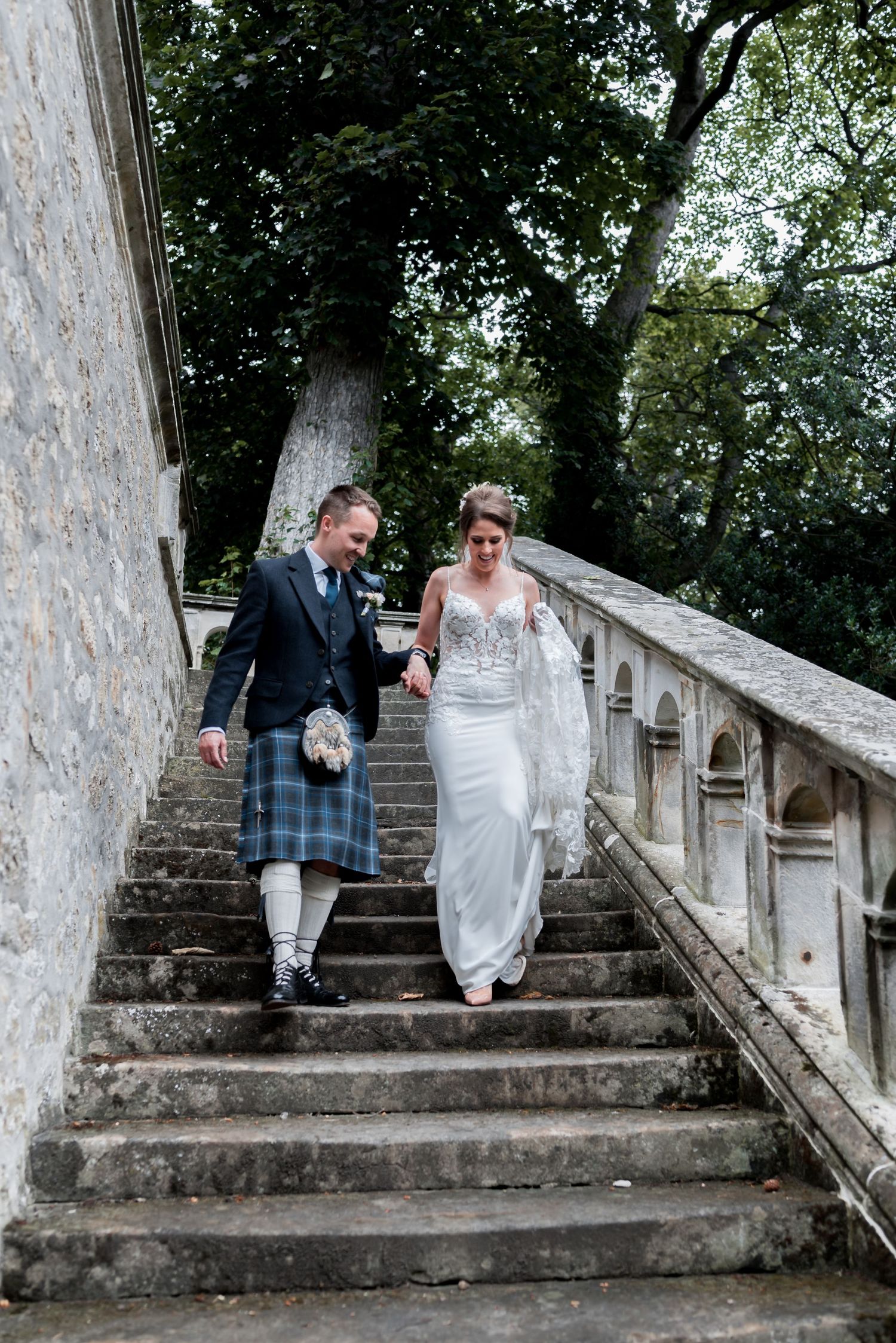 A couple in wedding attire walking down stone stairs outdoors, with the groom wearing traditional Scottish kilt attire.