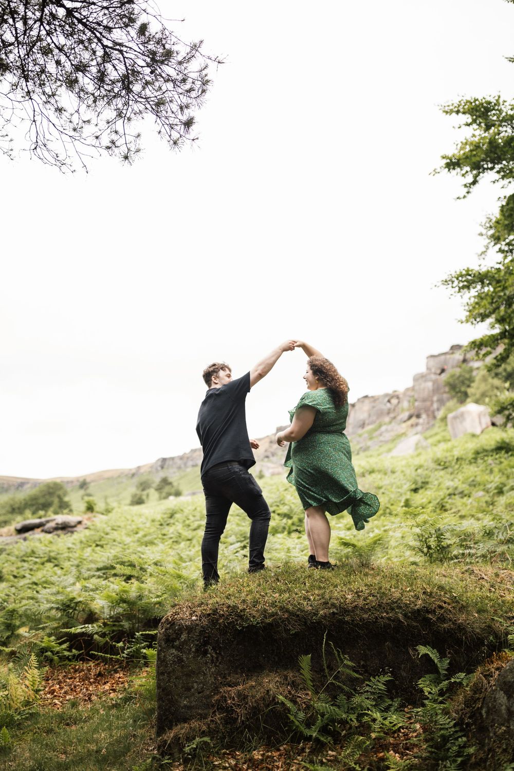 A couple dances joyfully on a grassy hilltop against a backdrop of mountains and trees.