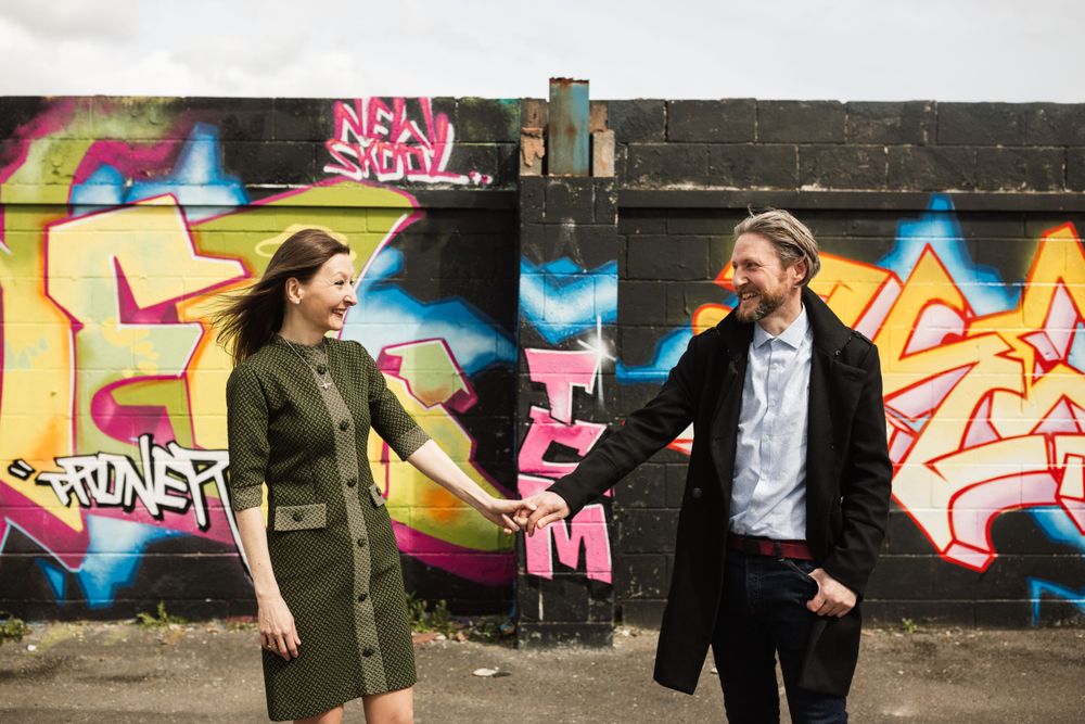 A stylish couple poses playfully in front of vibrant street art murals.