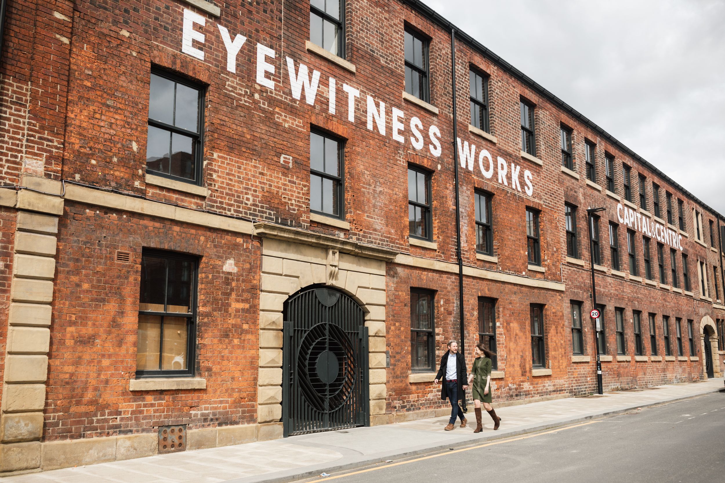 Couple walking past historic Eyewitness Works building with red brick facade.
