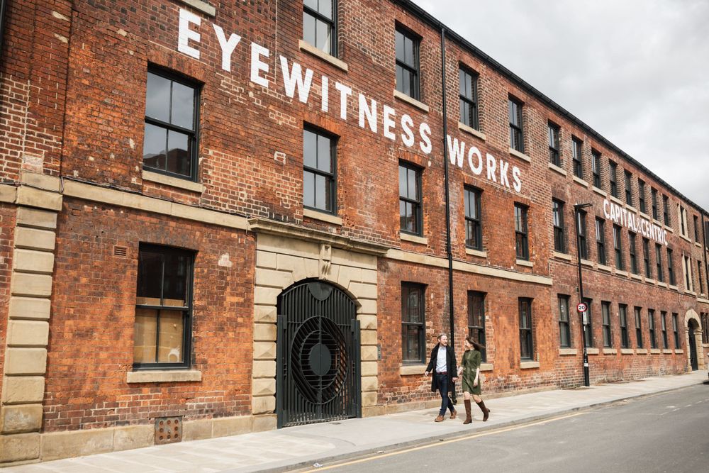 Couple walking past historic Eyewitness Works building with red brick facade.
