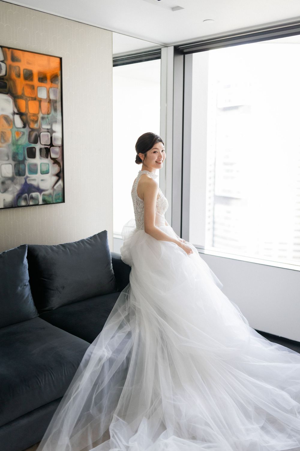 Bride in elegant white tulle ball gown smiles by large window with city views.