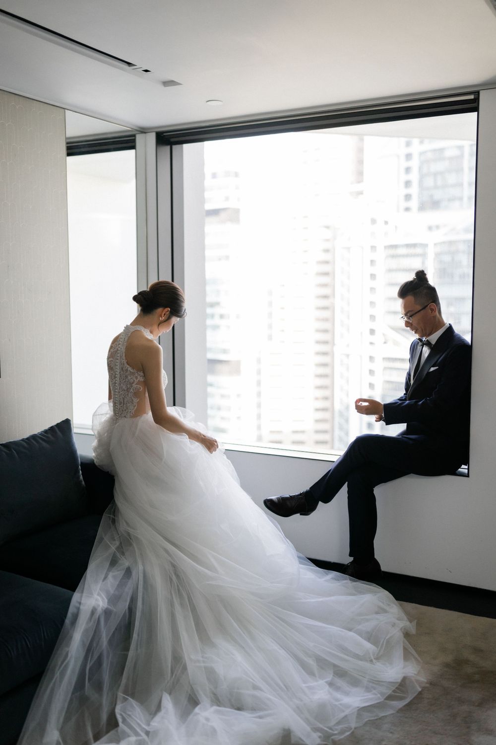 Bride and groom share quiet moment by floor-to-ceiling windows with city backdrop.