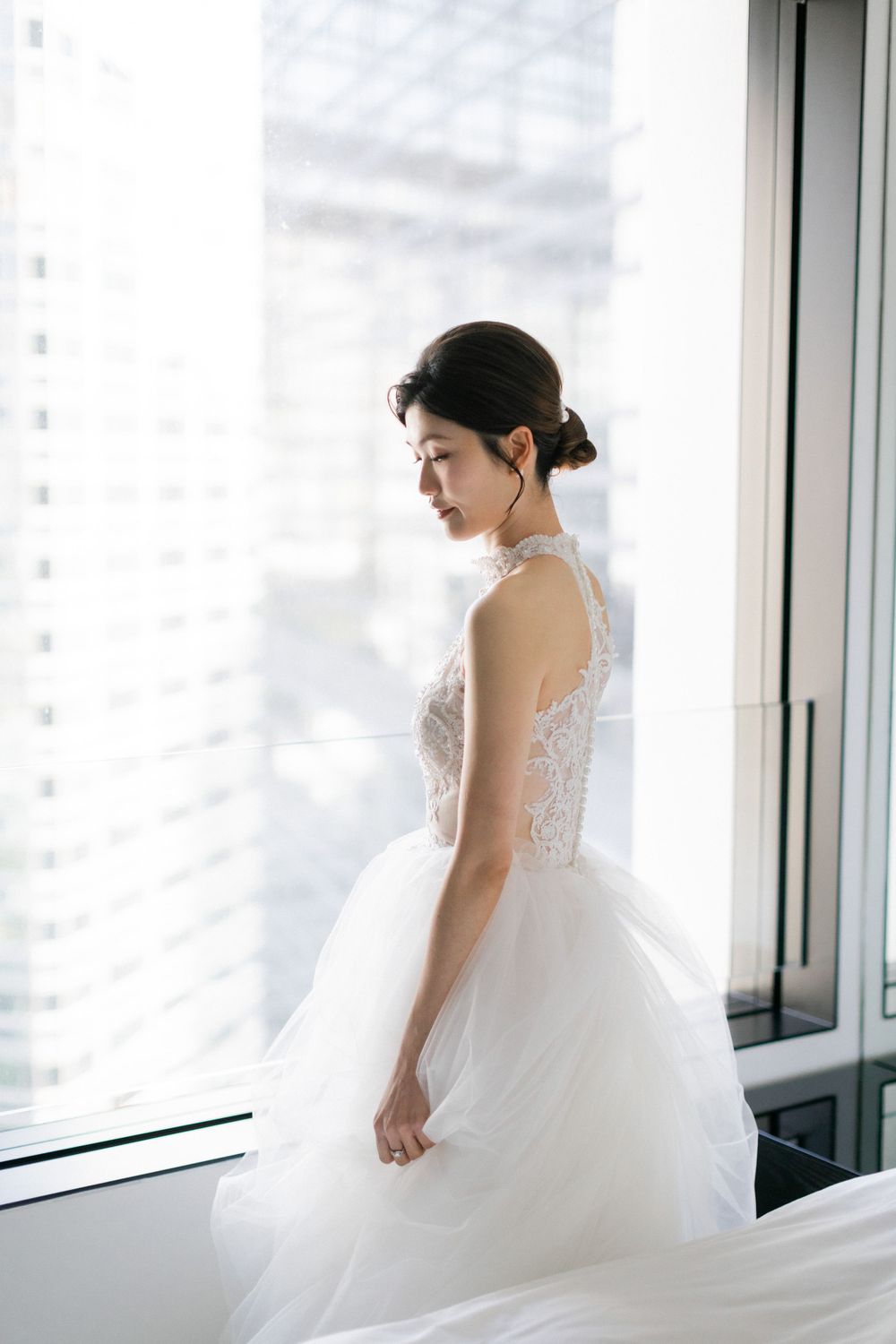 Elegant bride in white lace ballgown standing gracefully by a large city-view window.
