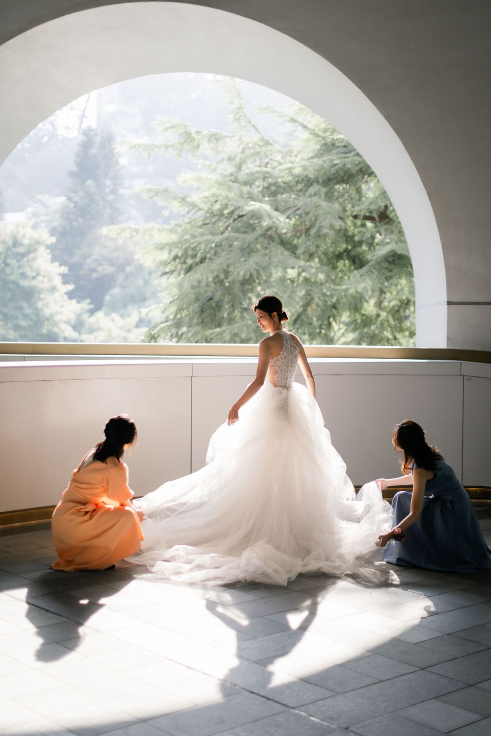 Two bridesmaids adjust bride's flowing white gown train in sunlit arched hallway.