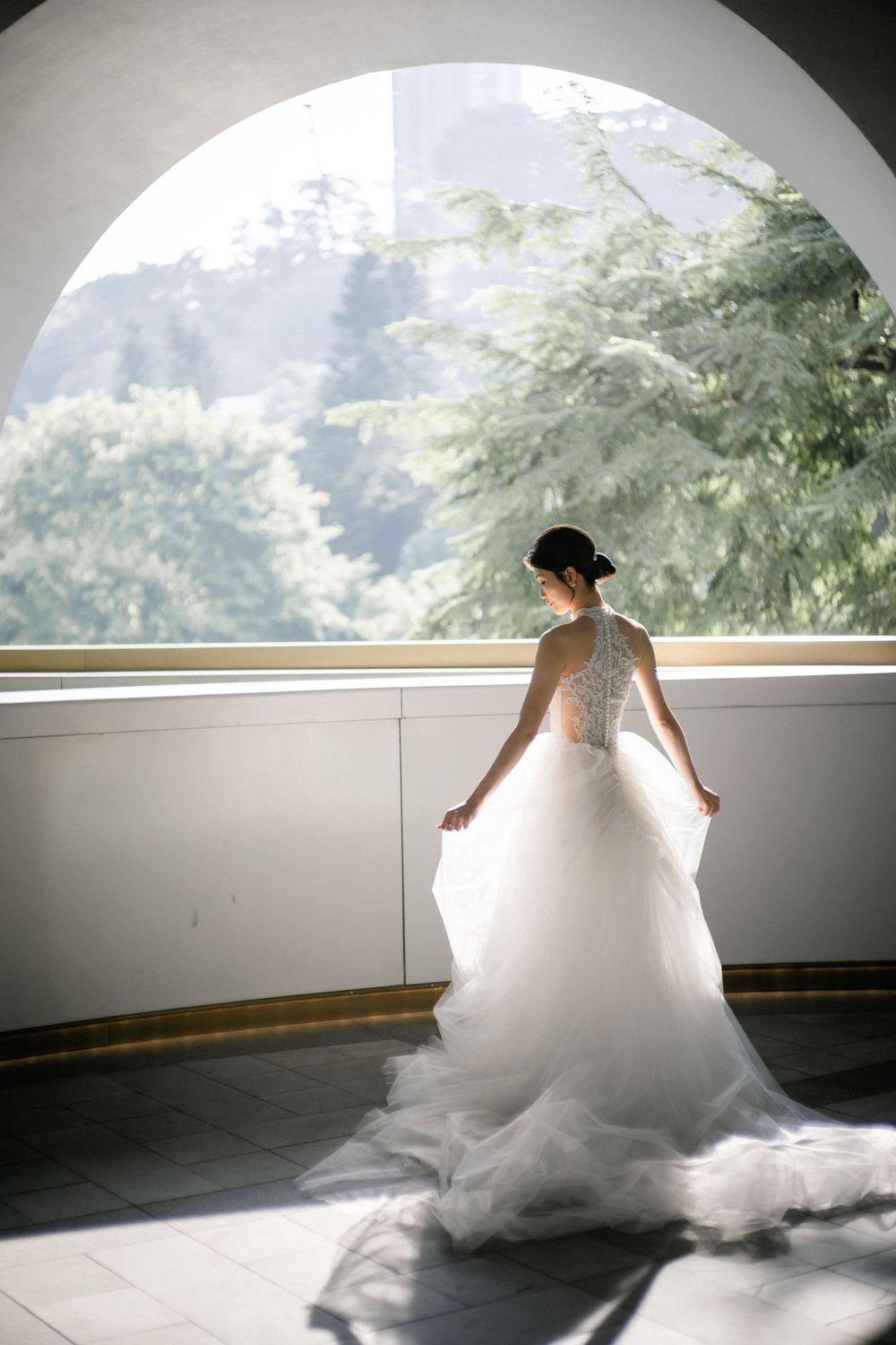 Bride in lace halter wedding gown poses gracefully in sunlit arched window alcove.