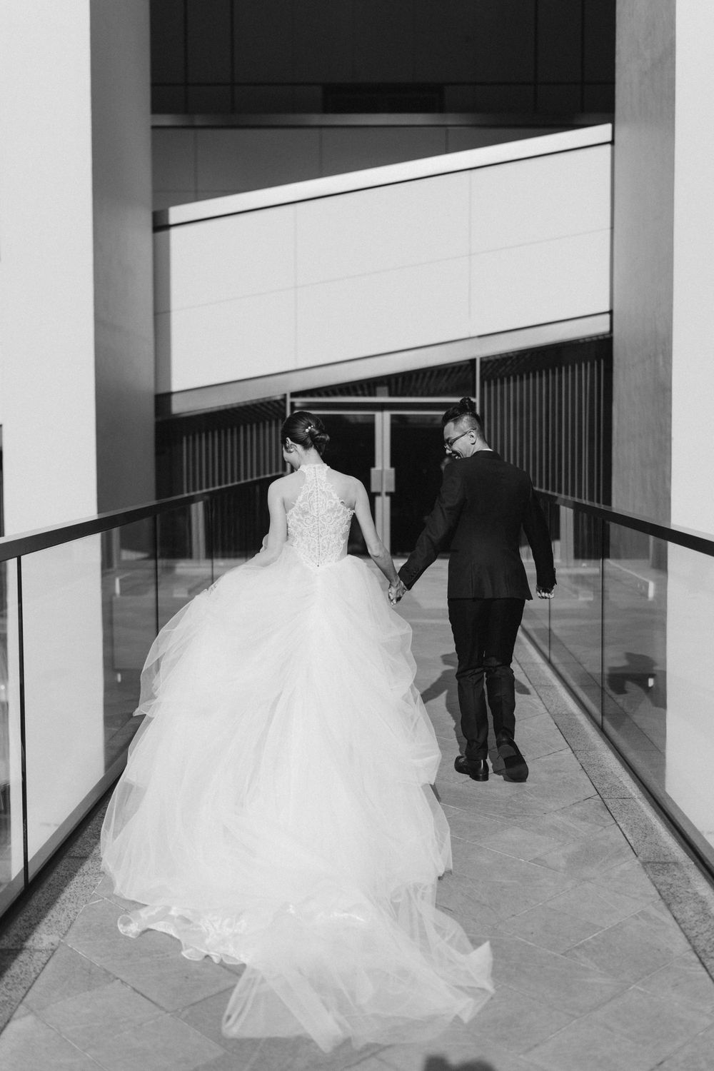 Black and white photo of bride and groom walking hand in hand down modern venue corridor.