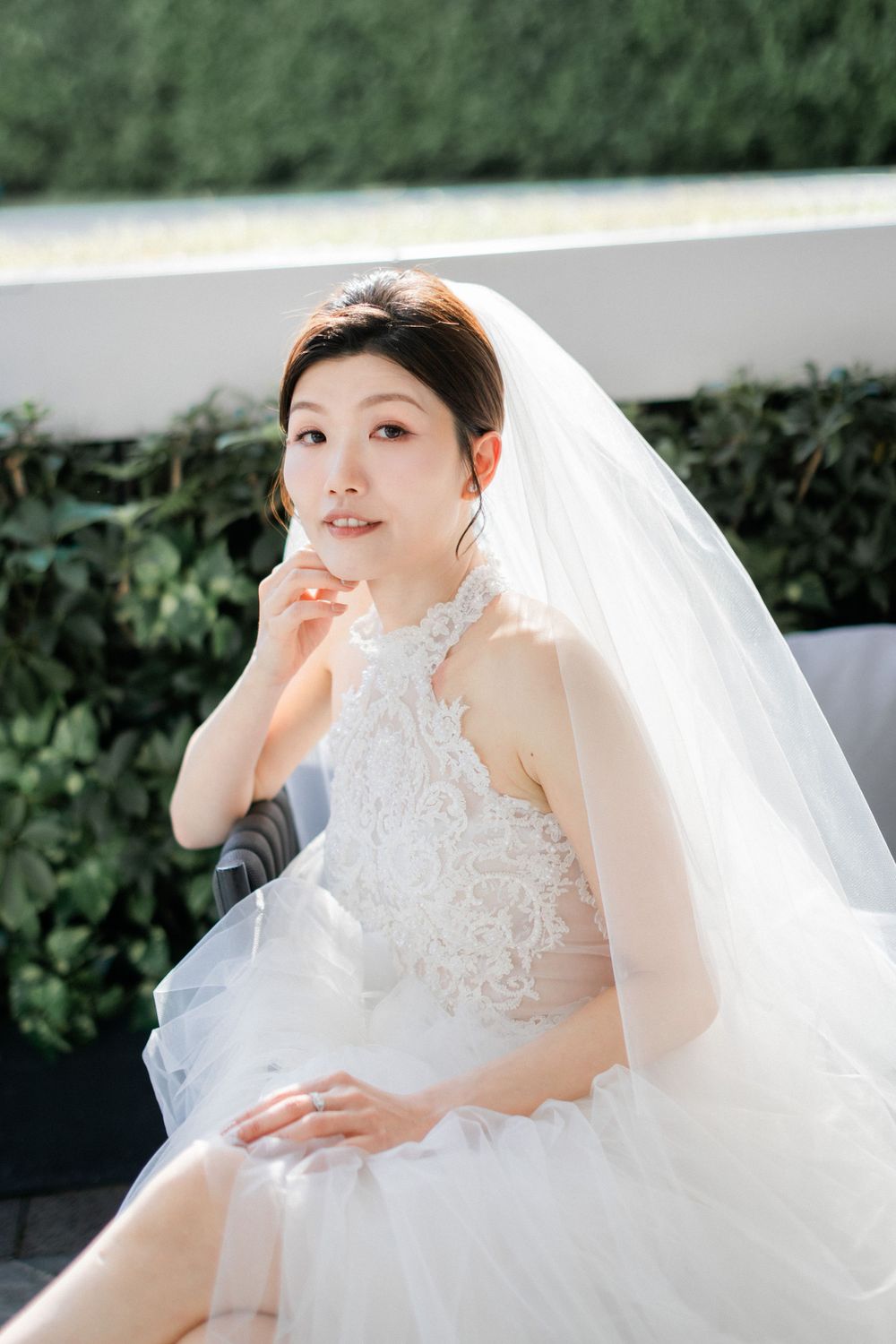 Elegant bride in white lace halter wedding gown and veil seated outdoors with greenery backdrop.
