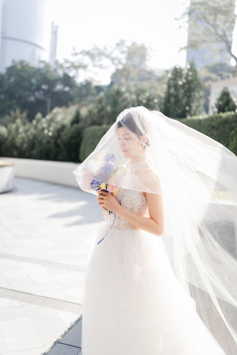 Bride in flowing white gown holding purple bouquet with veil billowing in sunlit outdoor setting.