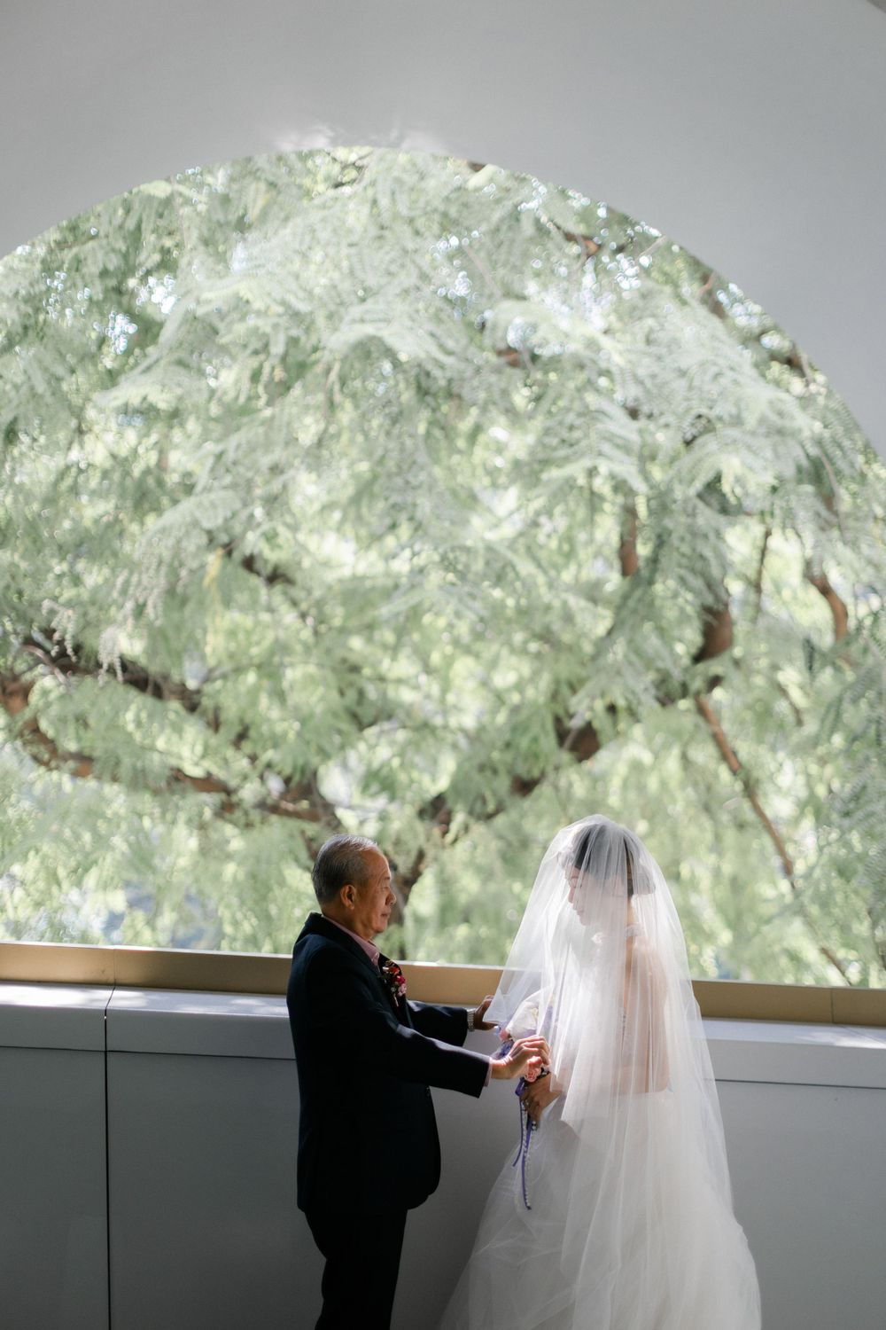 Bride and older gentleman sharing tender moment by large arched window with lush tree view.