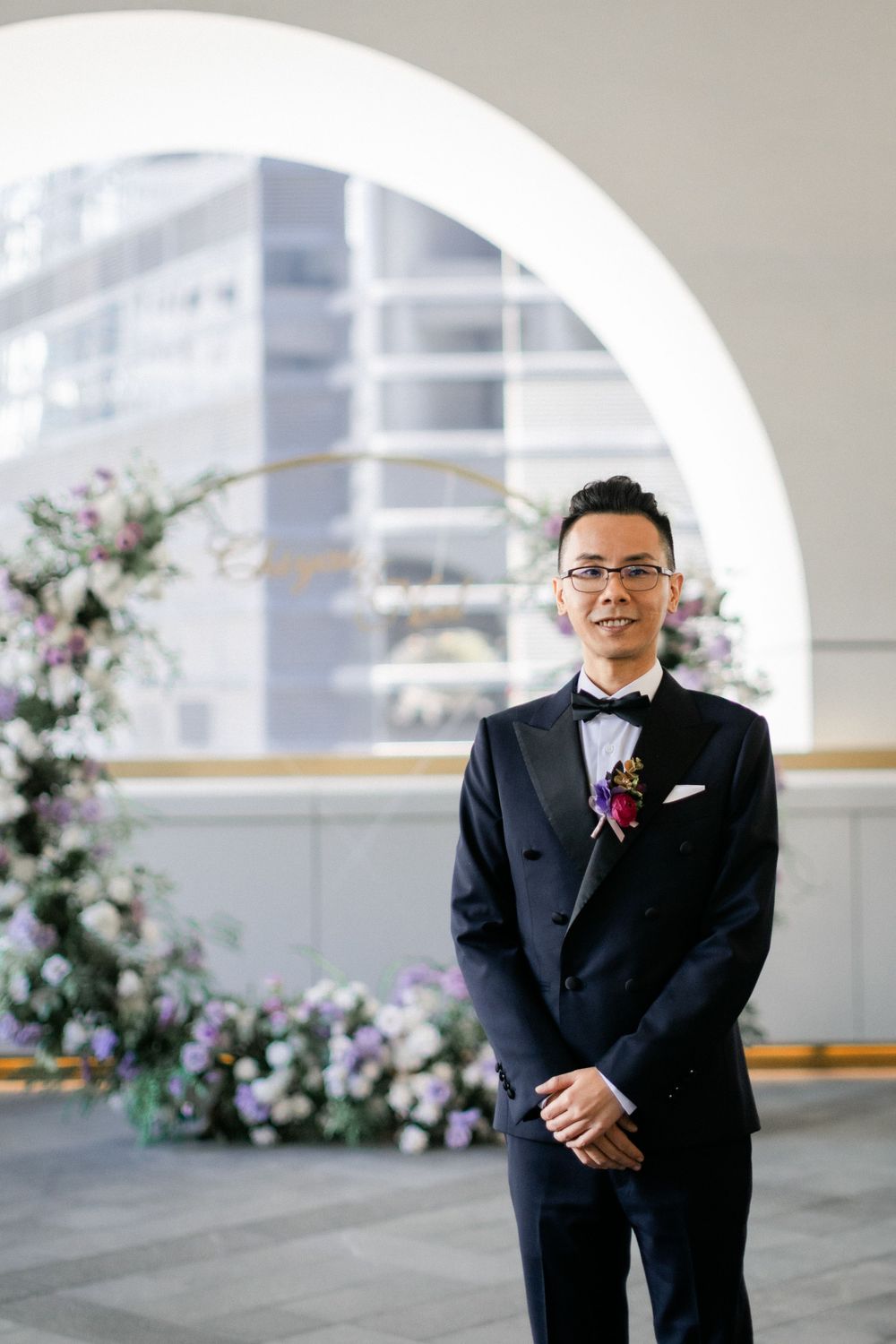 Dapper groom in black tuxedo with boutonniere standing before floral arch wedding ceremony backdrop.
