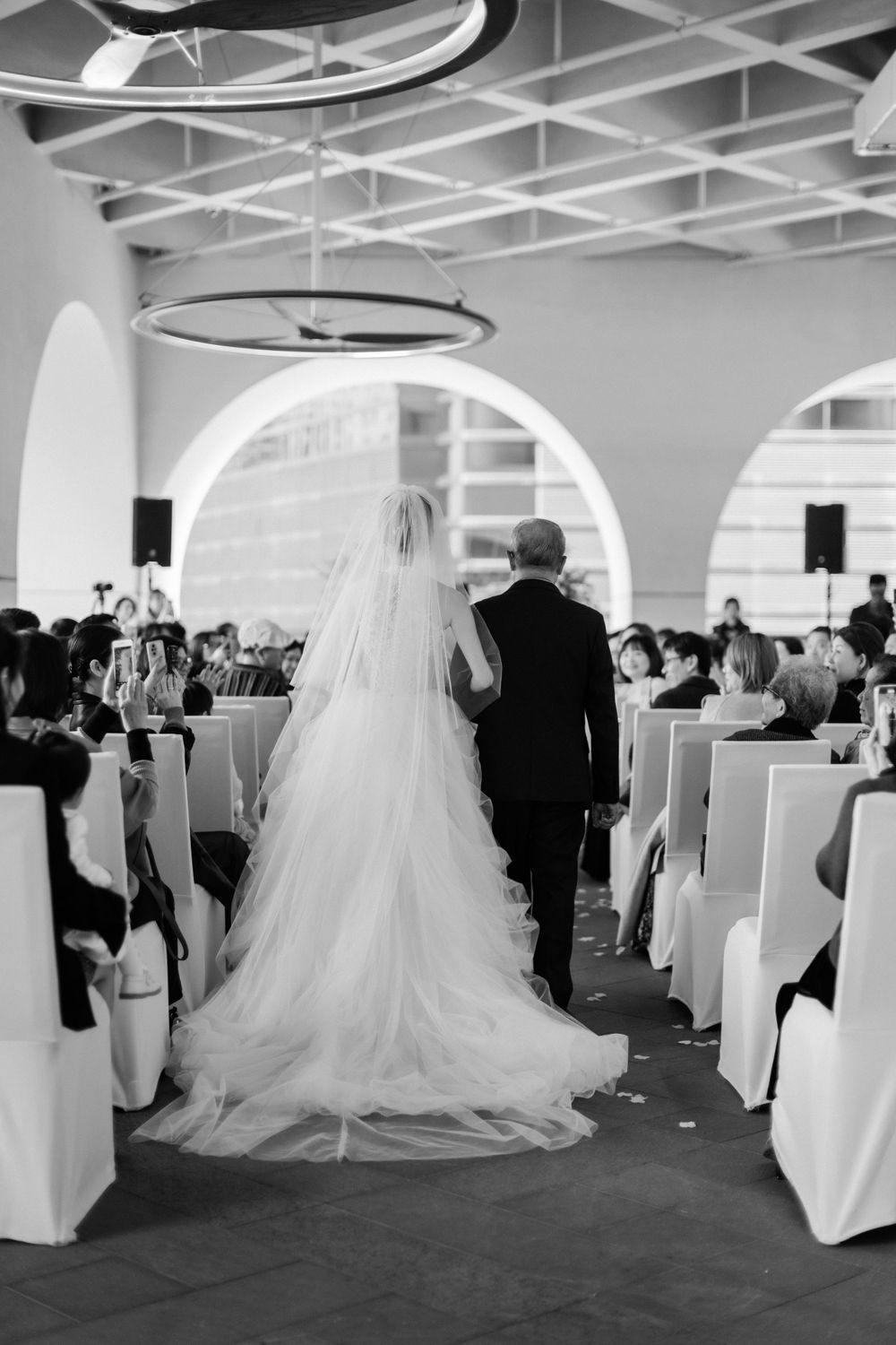 Black and white image of bride walking down aisle with escort during wedding ceremony.