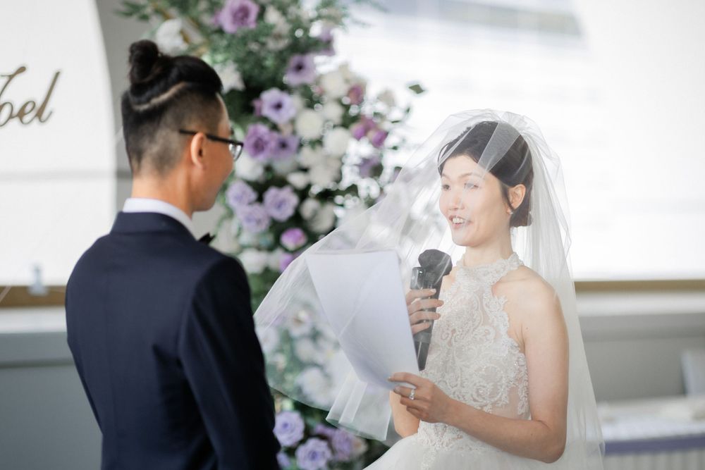 Bride reads wedding vows while holding microphone at elegant ceremony with purple floral backdrop.
