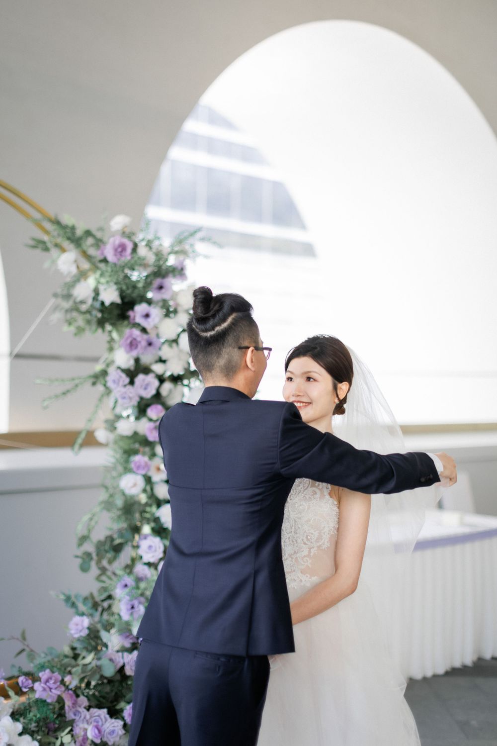 Bride and groom embrace near stunning purple and white floral arch at indoor wedding ceremony.