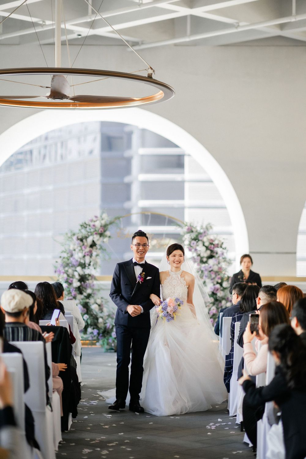 Smiling newlyweds walk down aisle together after ceremony in elegant venue with arched windows.