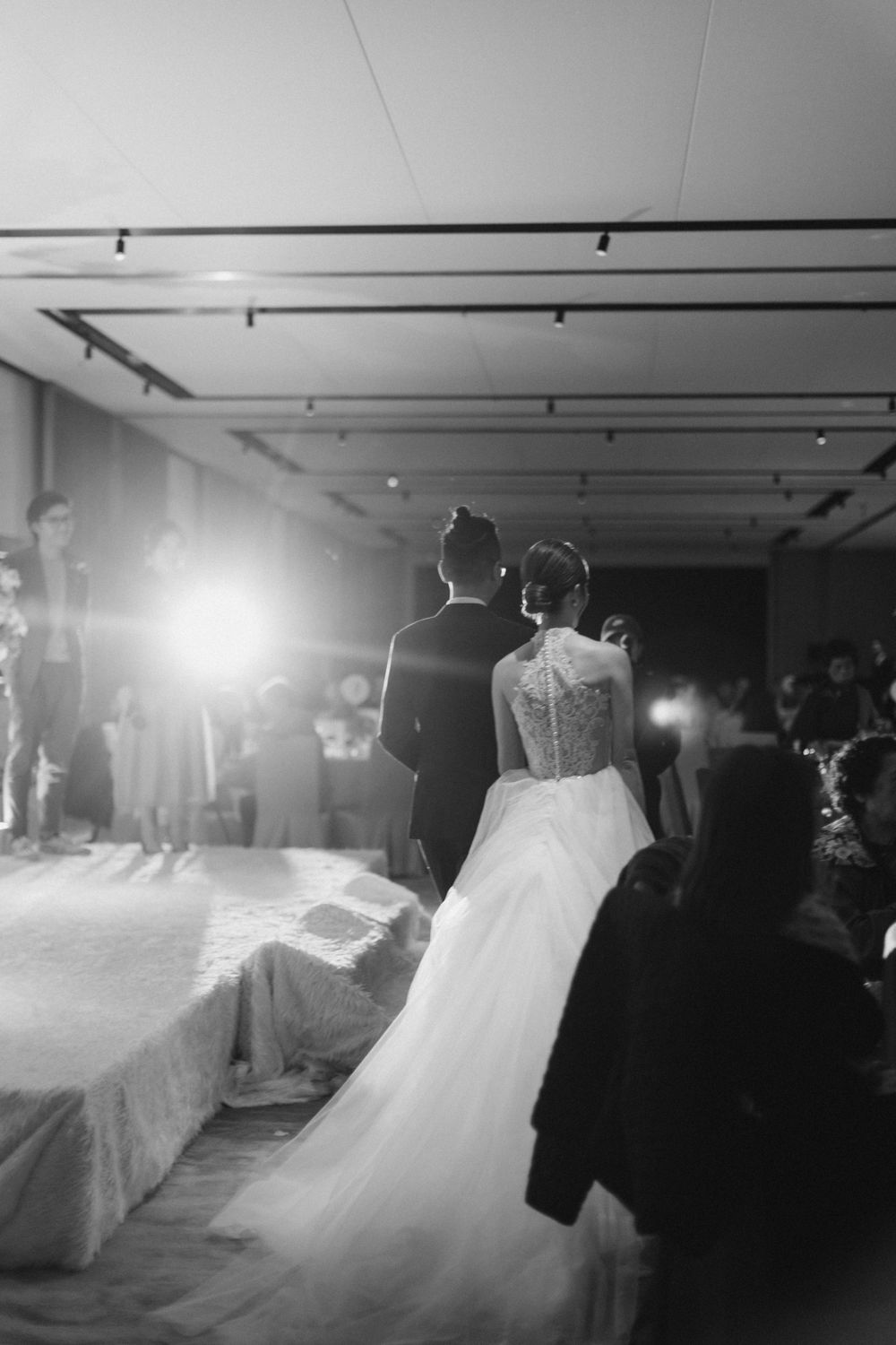 Bride and groom silhouetted against bright lights as they enter wedding reception in black and white.