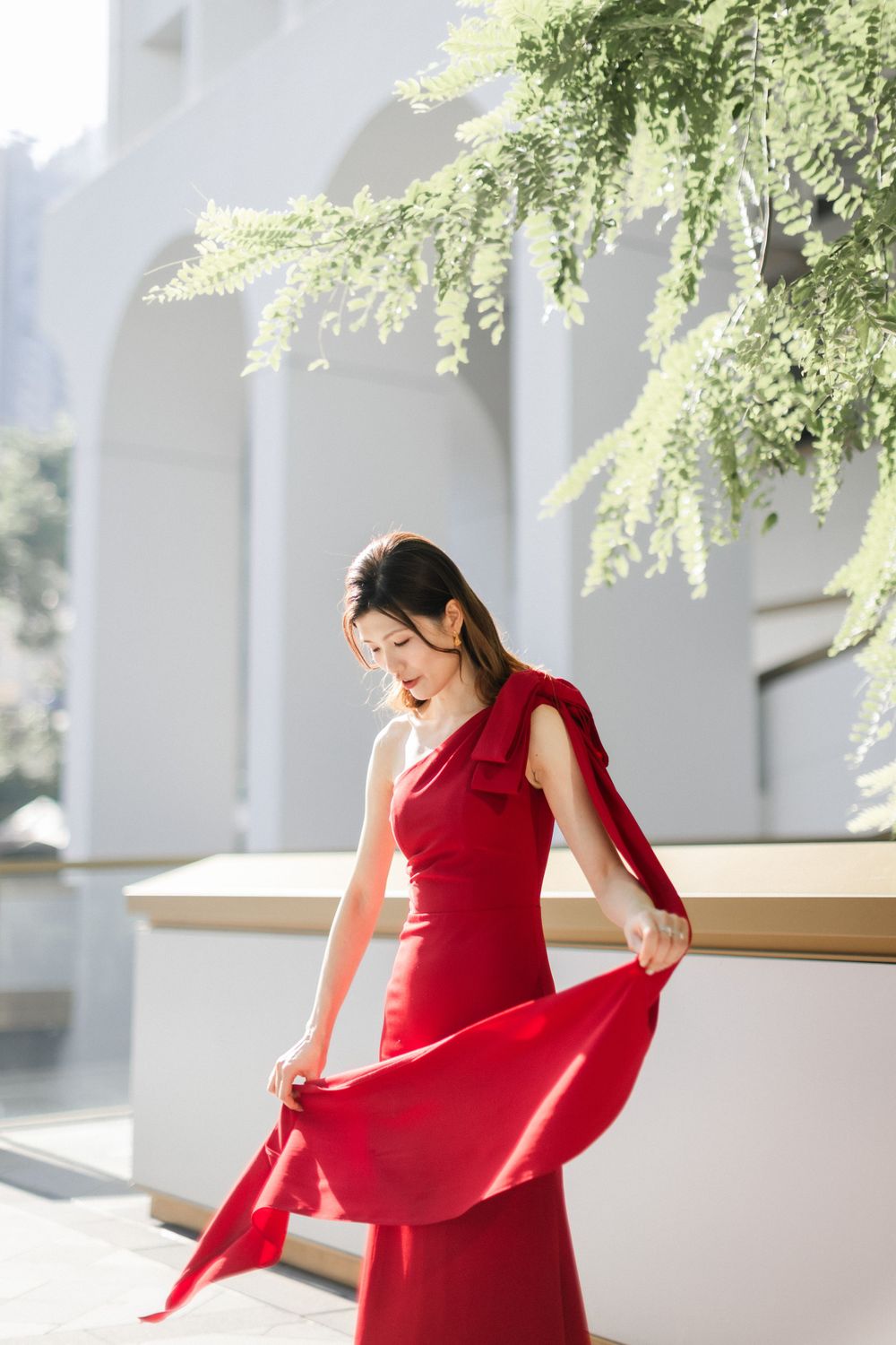 Elegant woman in red one-shoulder gown poses outdoors near white arched building in sunlight.