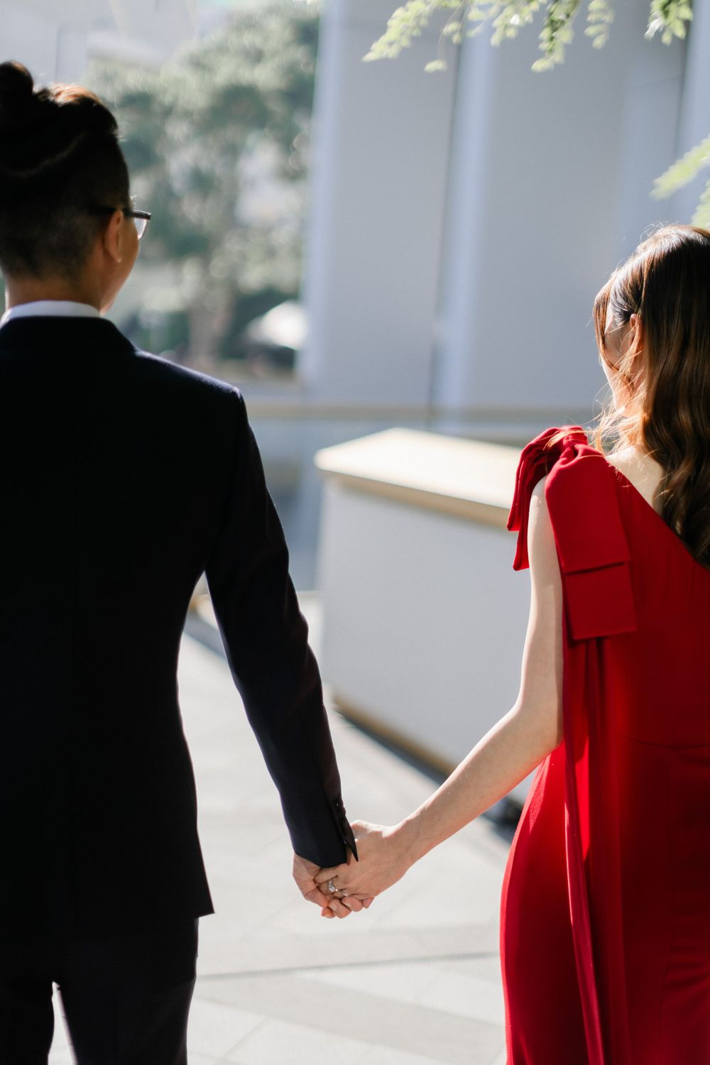 Couple holds hands walking away in sunlit outdoor setting, bride in flowing red gown.