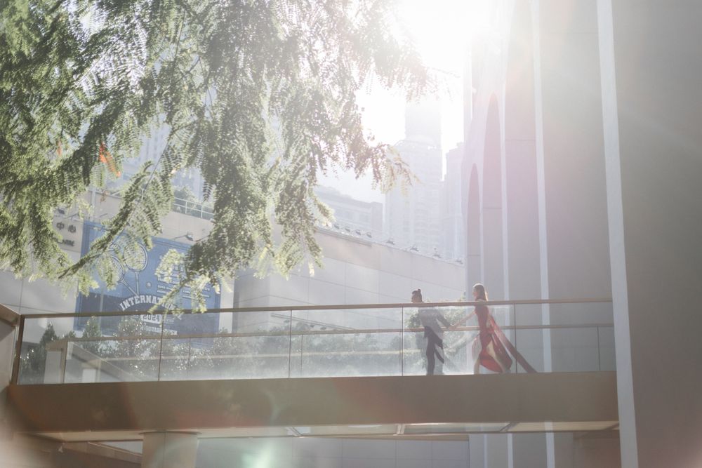 Couple strolls along sunlit outdoor balcony surrounded by lush green trees and modern building.