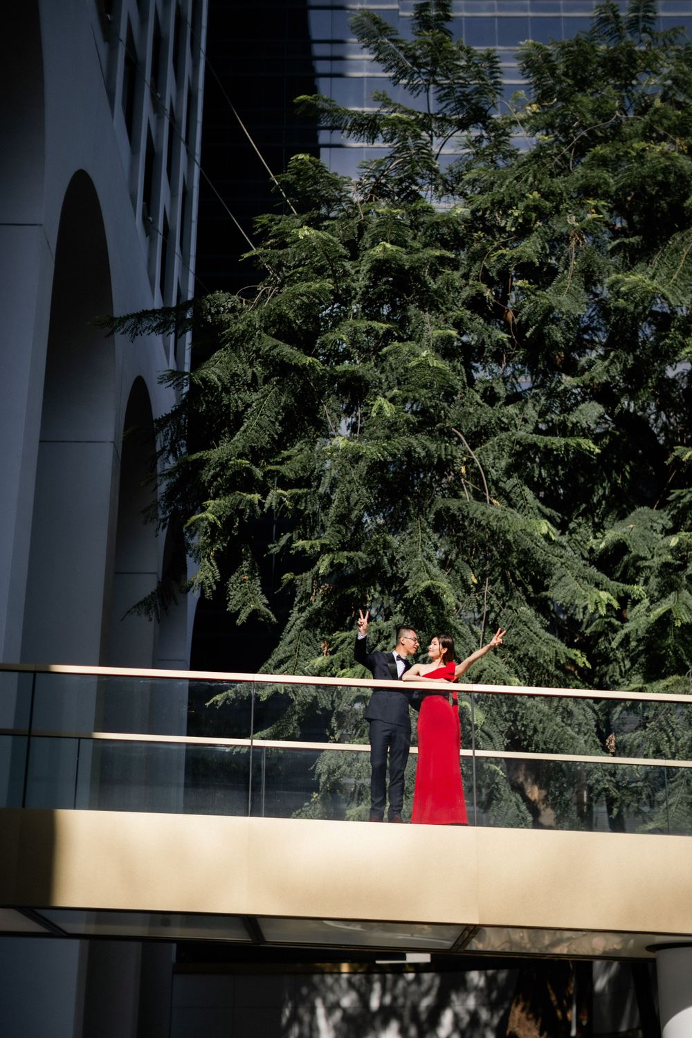 Couple in formal attire poses on golden balcony with arms outstretched amid lush green trees.