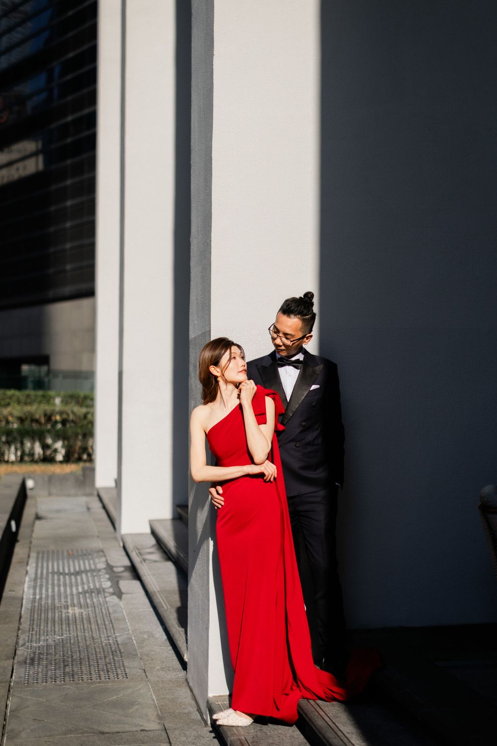 Couple in elegant attire pose against white wall, woman in red gown, man in black tuxedo.