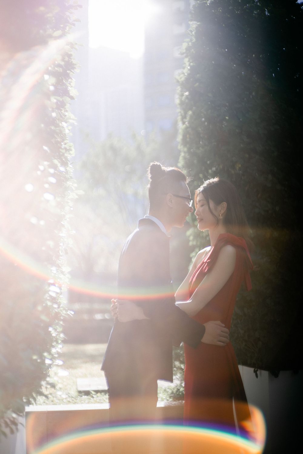 Romantic couple embrace in soft sunlight with lens flare, woman in red dress outdoors.