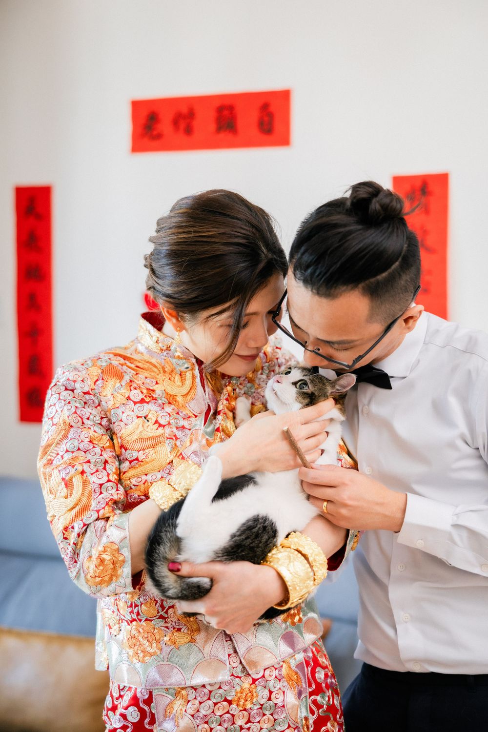 Newlywed couple in traditional Chinese wedding attire lovingly holding a cat with red banners behind.