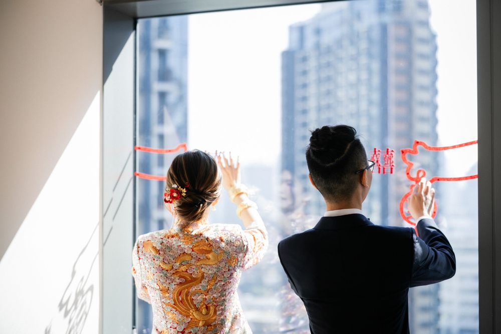 Bride and groom in traditional Chinese attire holding red wedding decorations by city-view window.