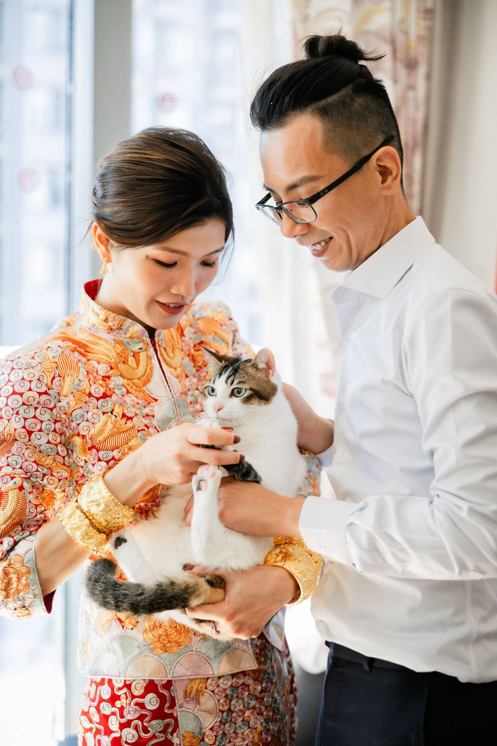 Smiling couple in Chinese wedding attire holding a calico cat near a bright window.