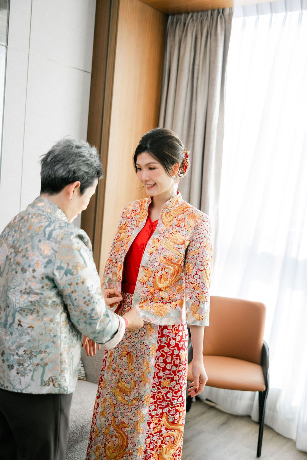 Bride in traditional Chinese qipao dress smiling as someone adjusts her colorful floral outfit indoors.